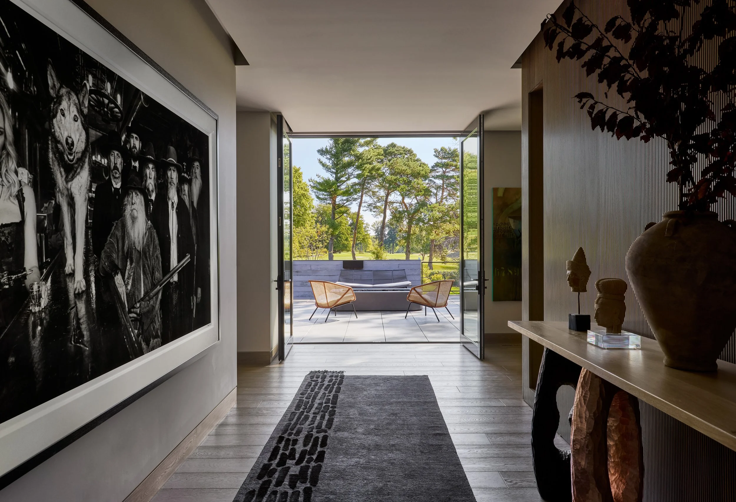 Interior of a modern home hallway leading to an outdoor patio with trees in view, decorated with artwork, sculptures, and furniture.