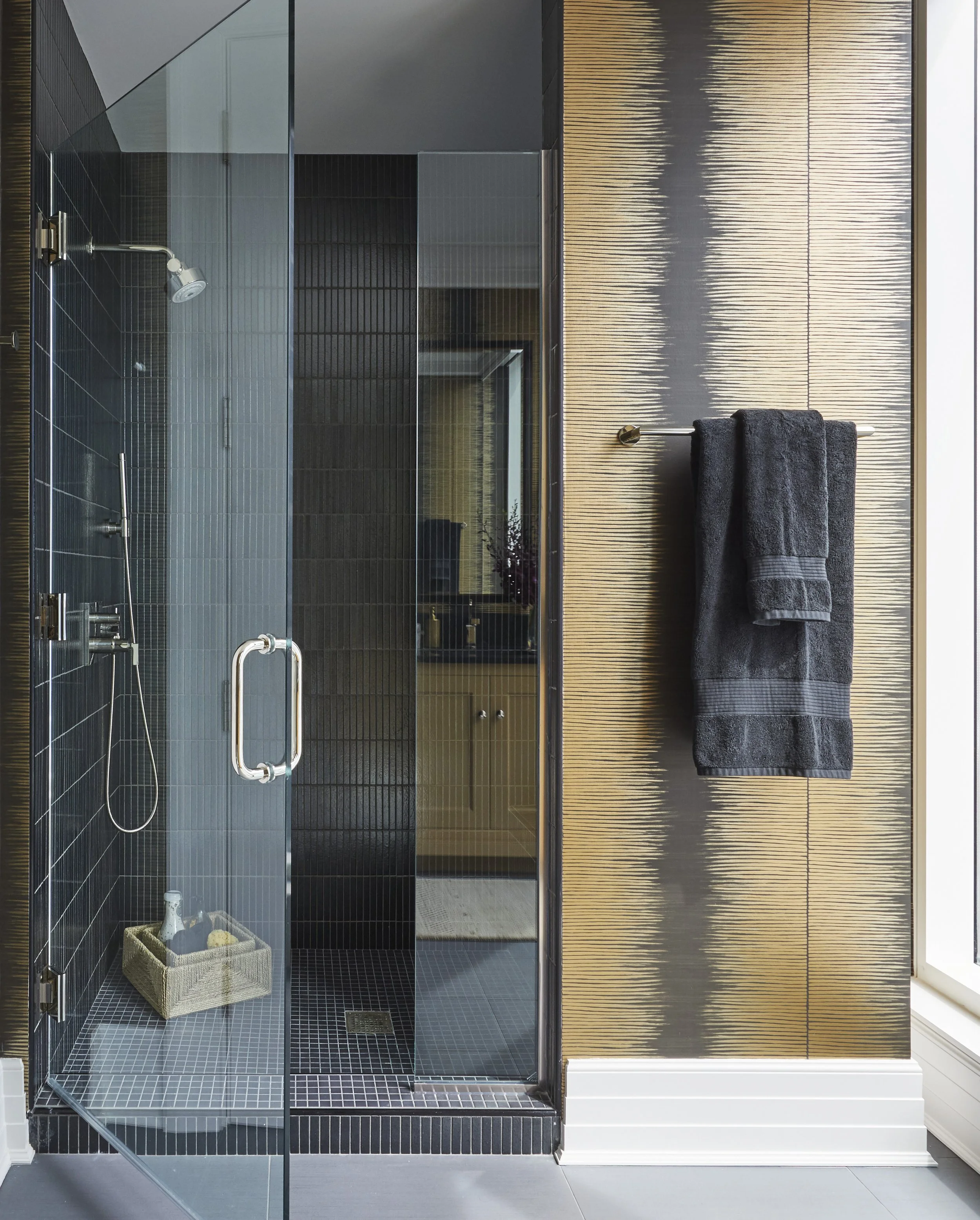 A modern, glass-enclosed walk-in shower with a black tile interior, a small basket with toiletries on the floor, and dark towels hanging on a metallic towel rod on a textured wooden wall adjacent to the shower.