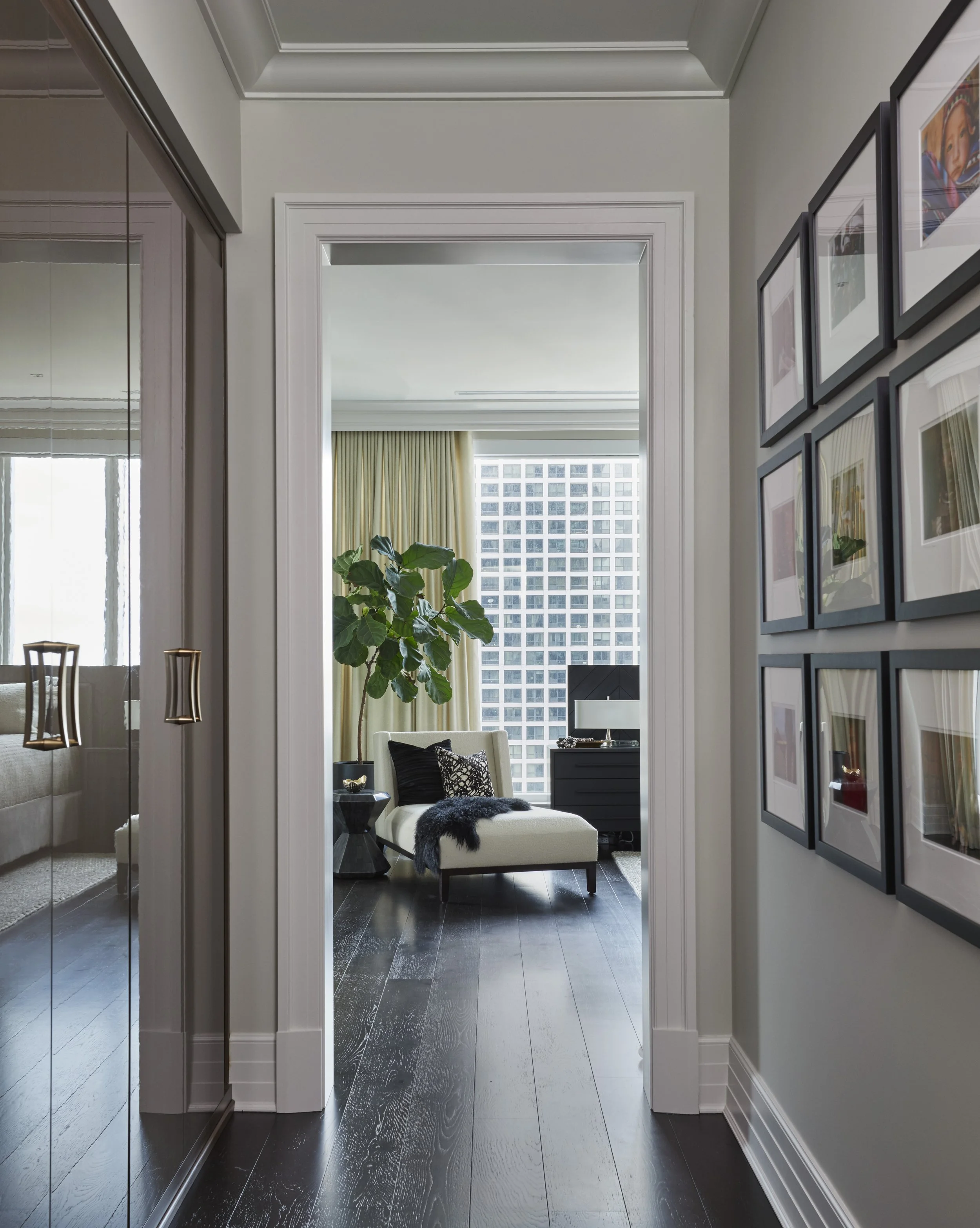 View through a hallway into a living room with large window, beige curtains, and a white ottoman with black and white pillows. Potted plant on side table. Framed photographs on wall to the right. Dark hardwood floor.