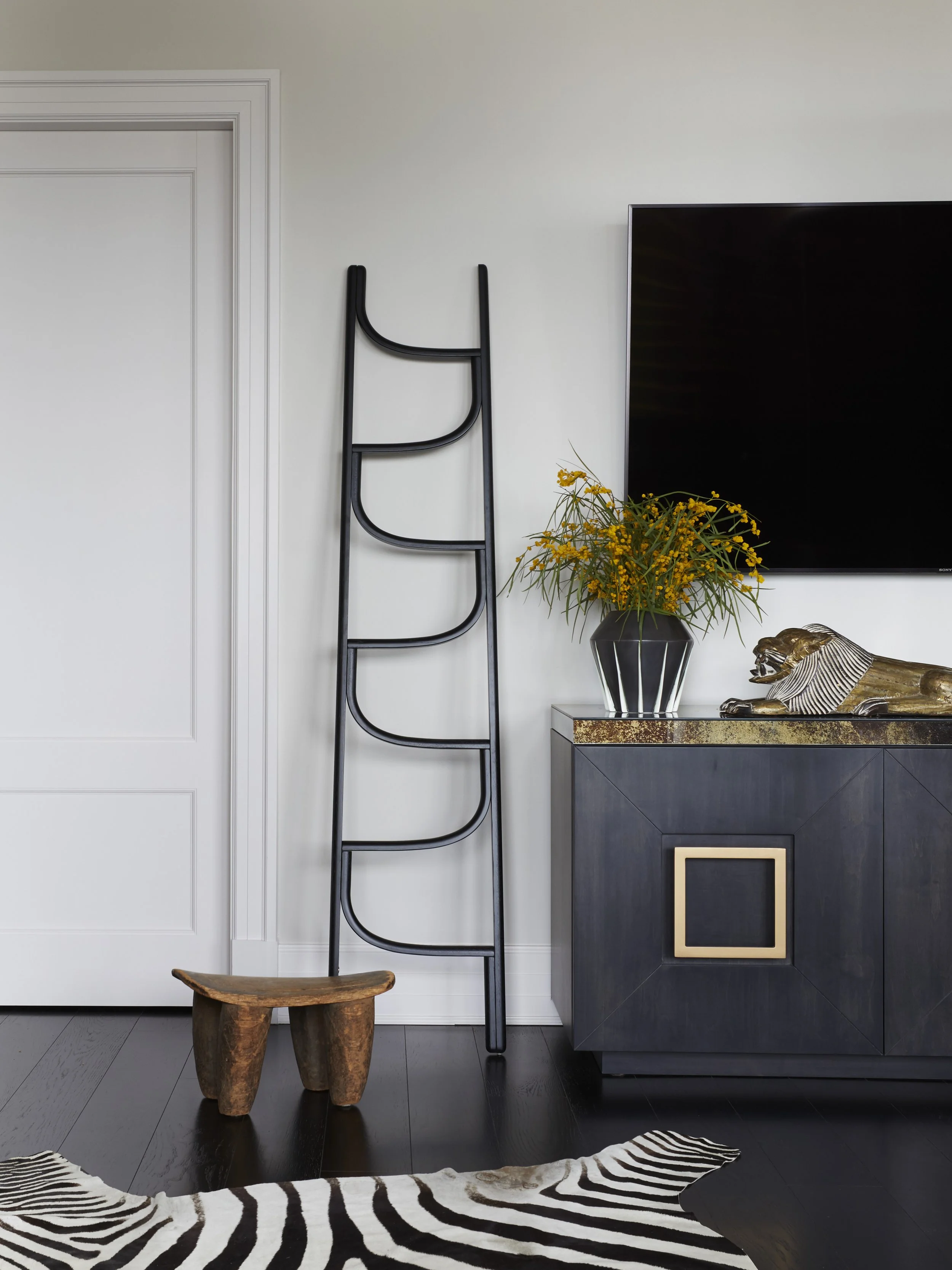 Living room with a white wall, a tall black ladder, a vintage black cabinet with a gold square handle, a vase of yellow flowers, a wooden stool, a living room rug with zebra pattern, a zebra print cushion, a gold decorative tiger sculpture, and a fla