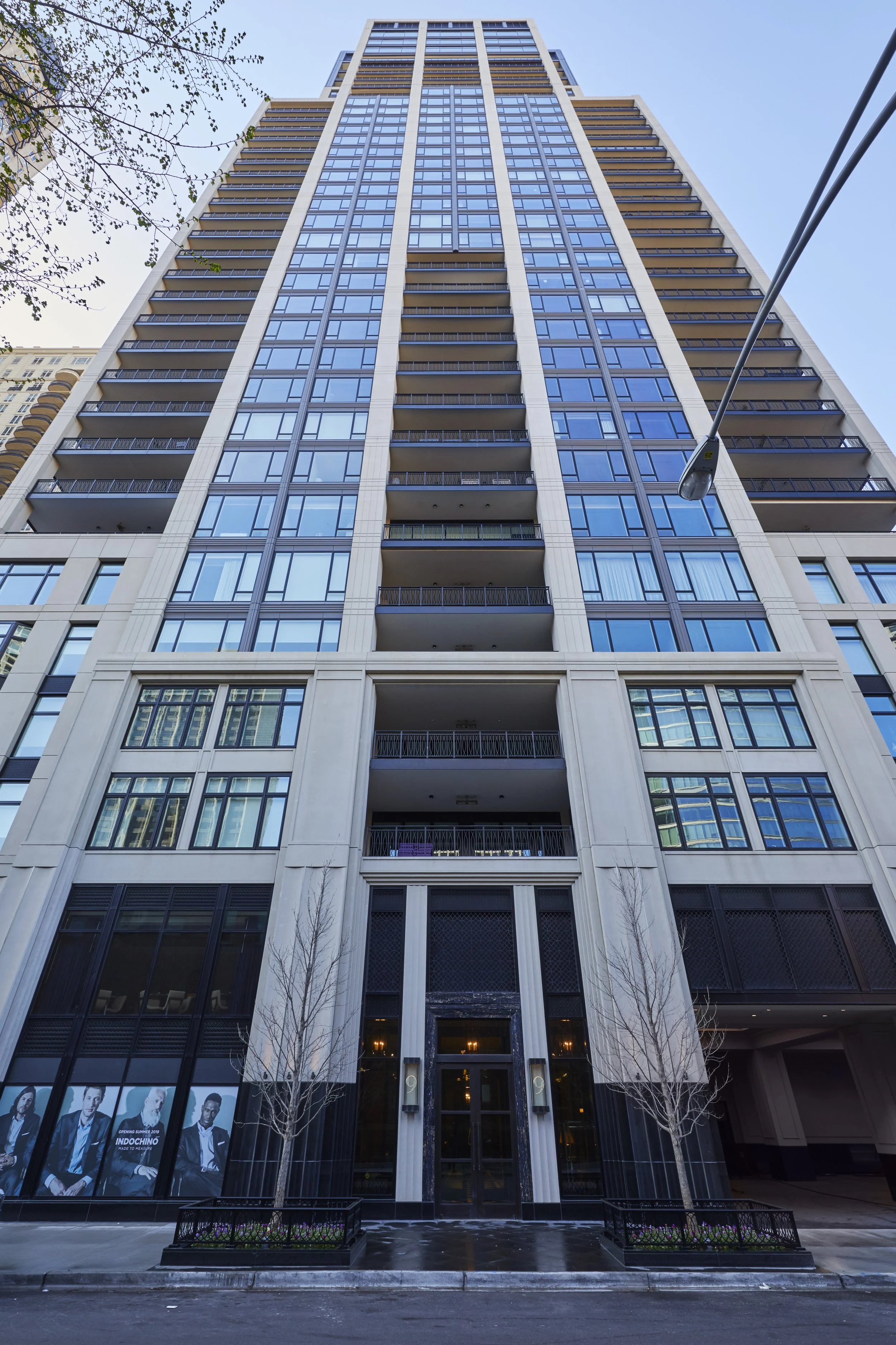 View of a tall modern building from ground level, with glass windows and balconies, and a street lamp in the foreground under a clear blue sky.