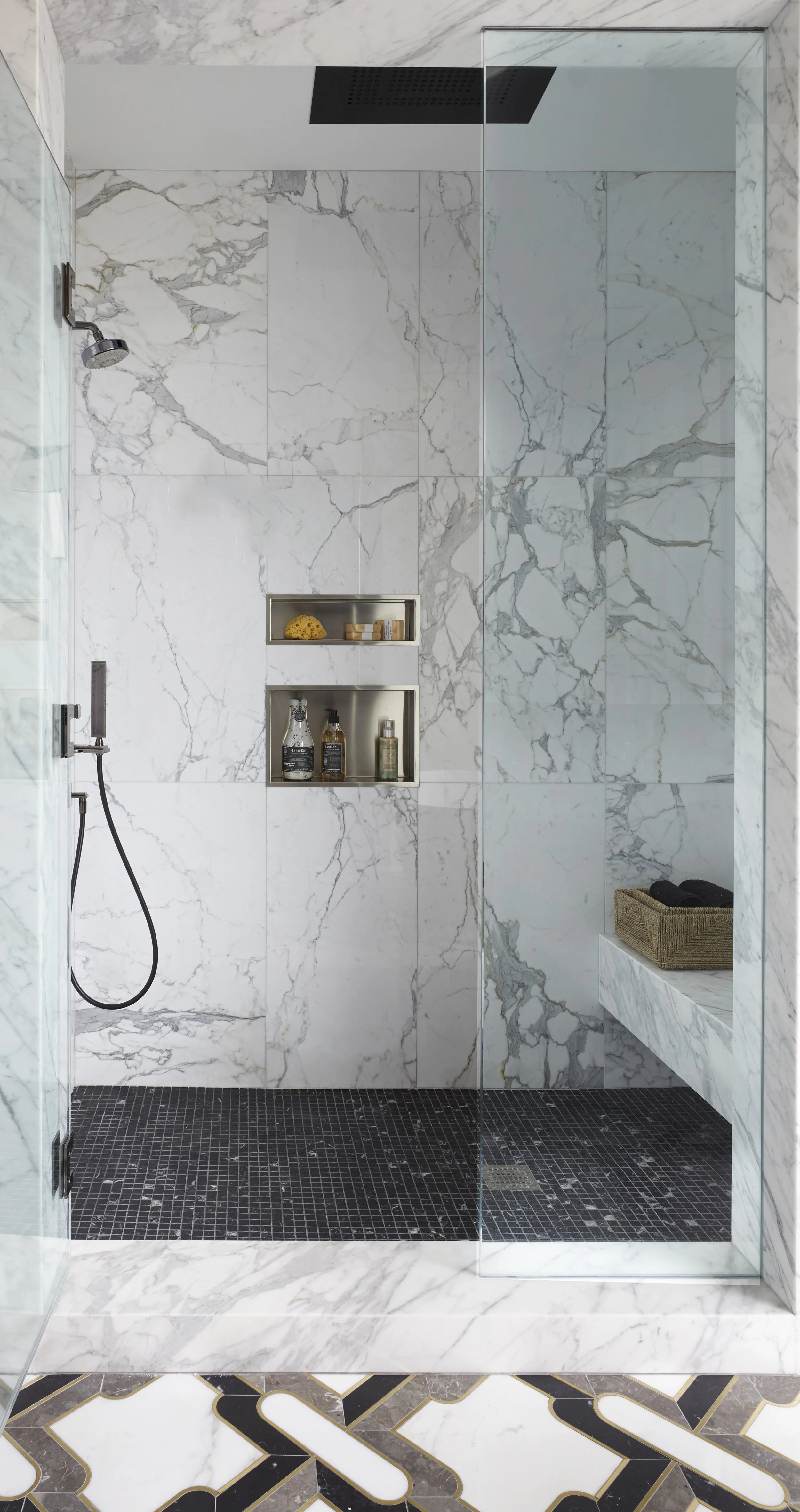 Modern marble shower with glass enclosure, black showerhead, built-in shelves with shampoo and soap, and a bench with towels.