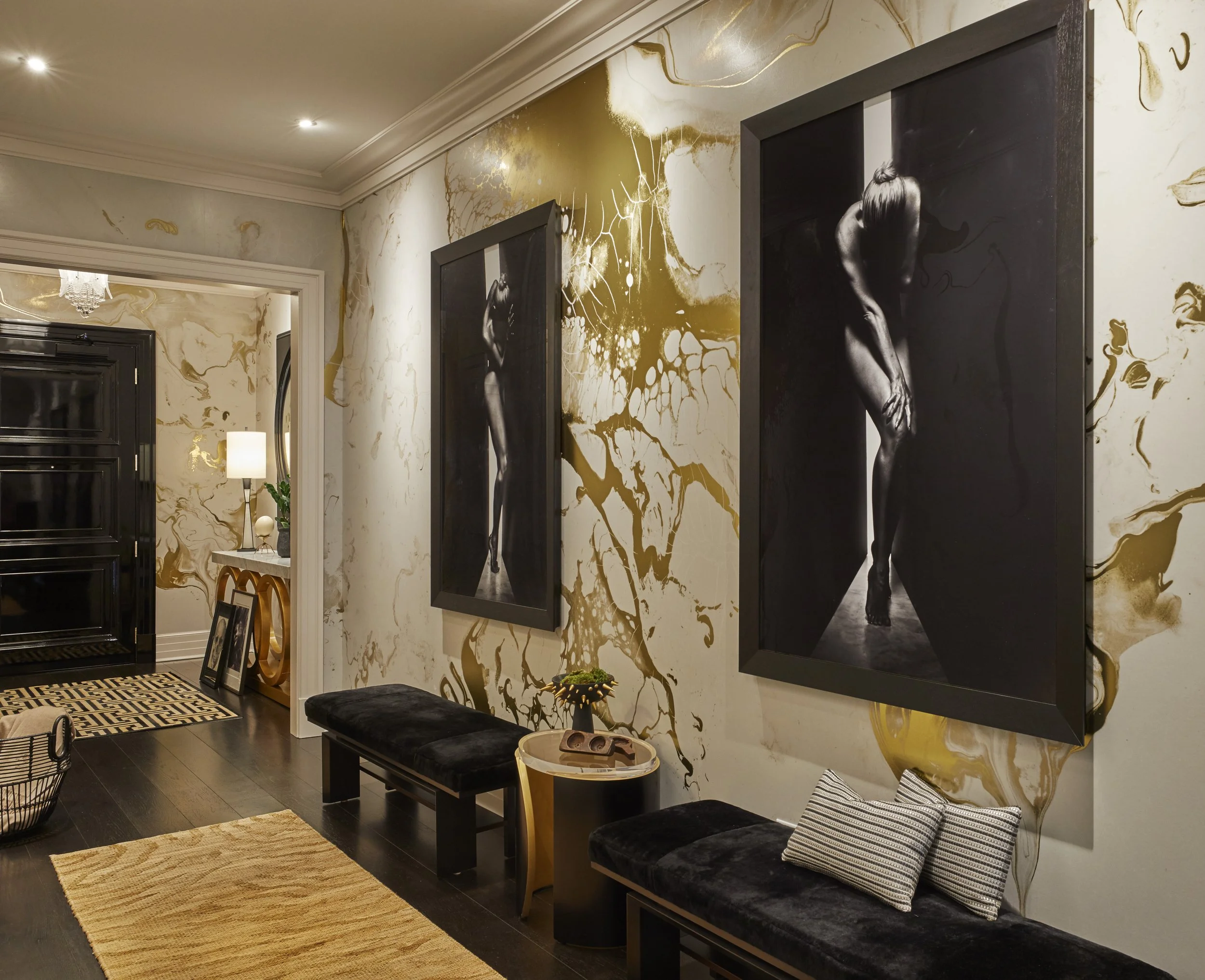 Living room with black and gold decor, black artwork of a woman on the wall, beige and gold marbled wallpaper, black bench with cushions, beige rug, and a small round table with plants and a decorative item.