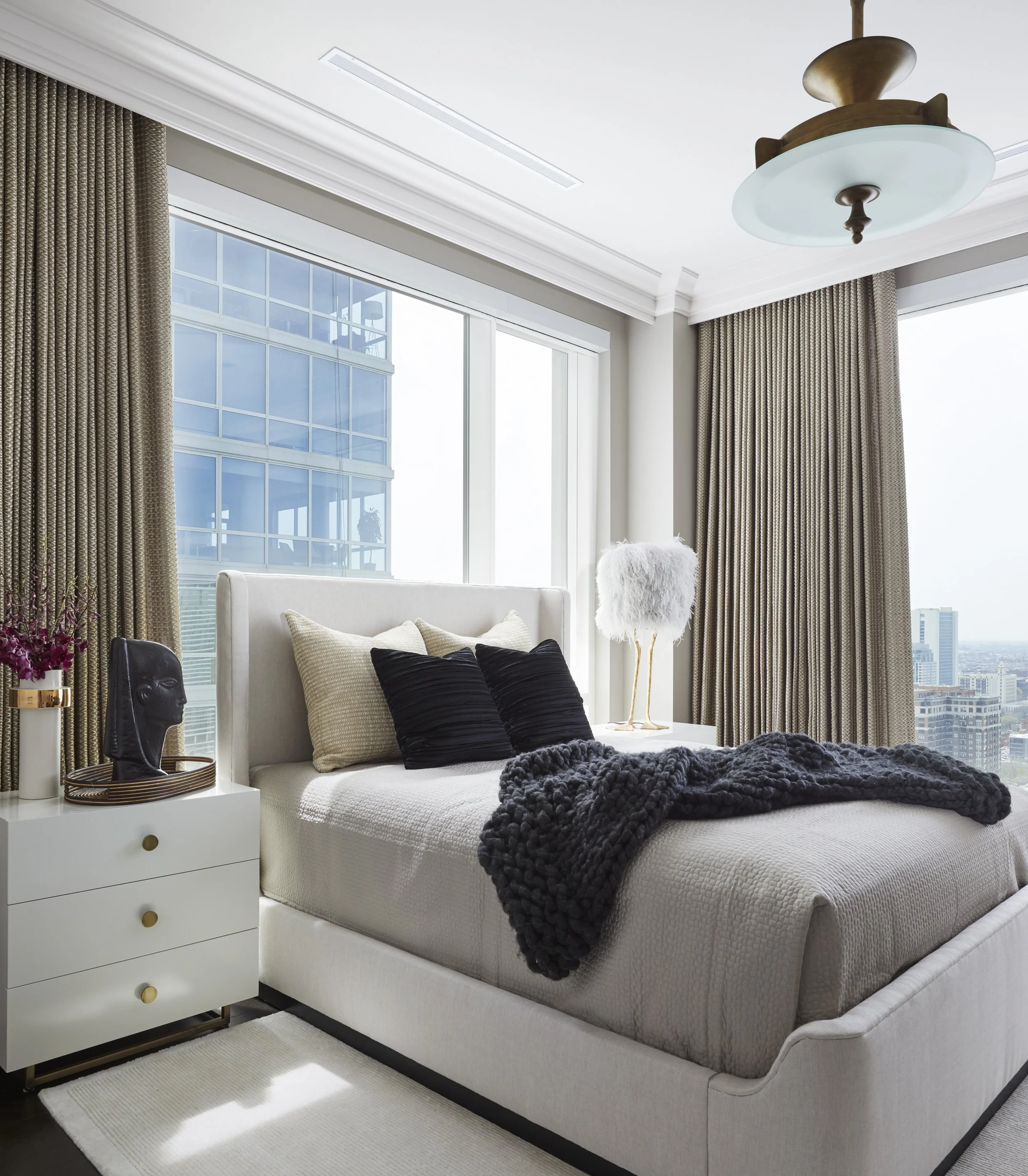 A modern bedroom with a white bed, black and beige pillows, a dark textured blanket, a white dresser with gold knobs, a decorative black face sculpture, a tall fluffy white lamp, beige curtains, large windows showing city skyscrapers, roman blinds, a