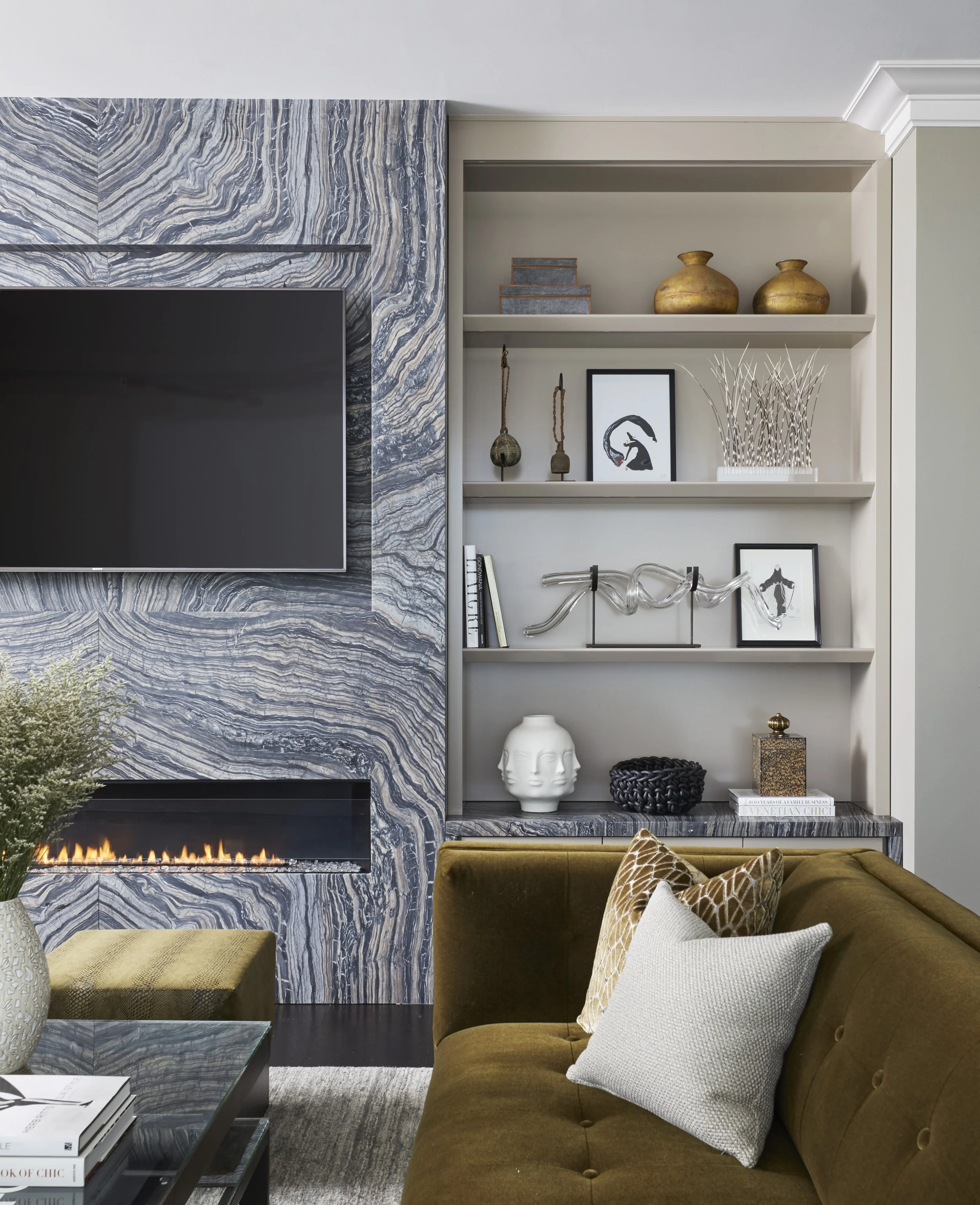 Living room with a marble fireplace and built-in shelves displaying vases, art, and decorative objects, with a brown velvet sofa and decorative pillows in the foreground.
