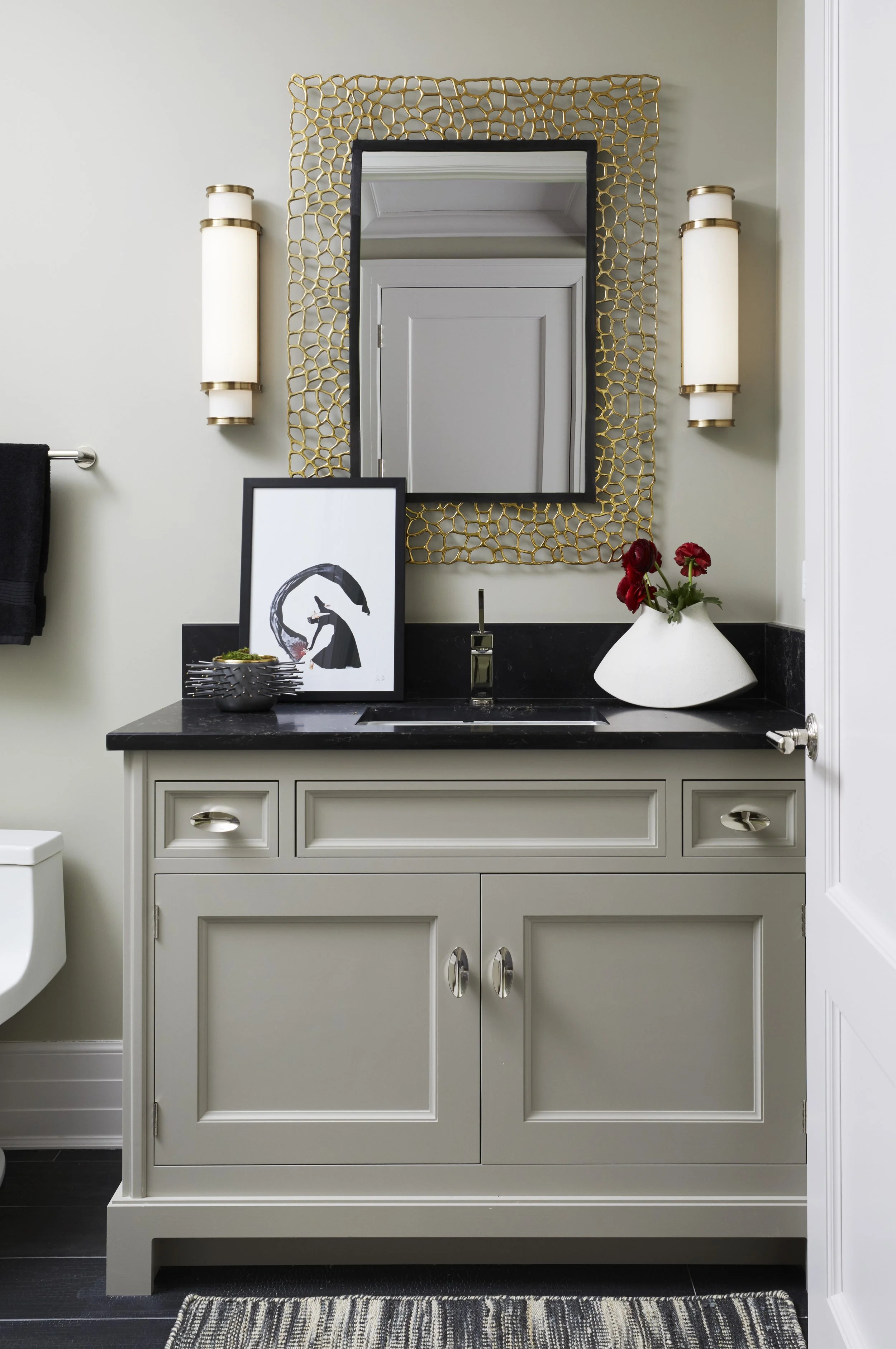 A bathroom vanity with a gray cabinet, black countertop, and a sink. On the countertop, there is a framed black-and-white artwork, a small bowl, and a white vase with red flowers. Above the vanity, a mirror with a gold honeycomb-patterned frame is mo