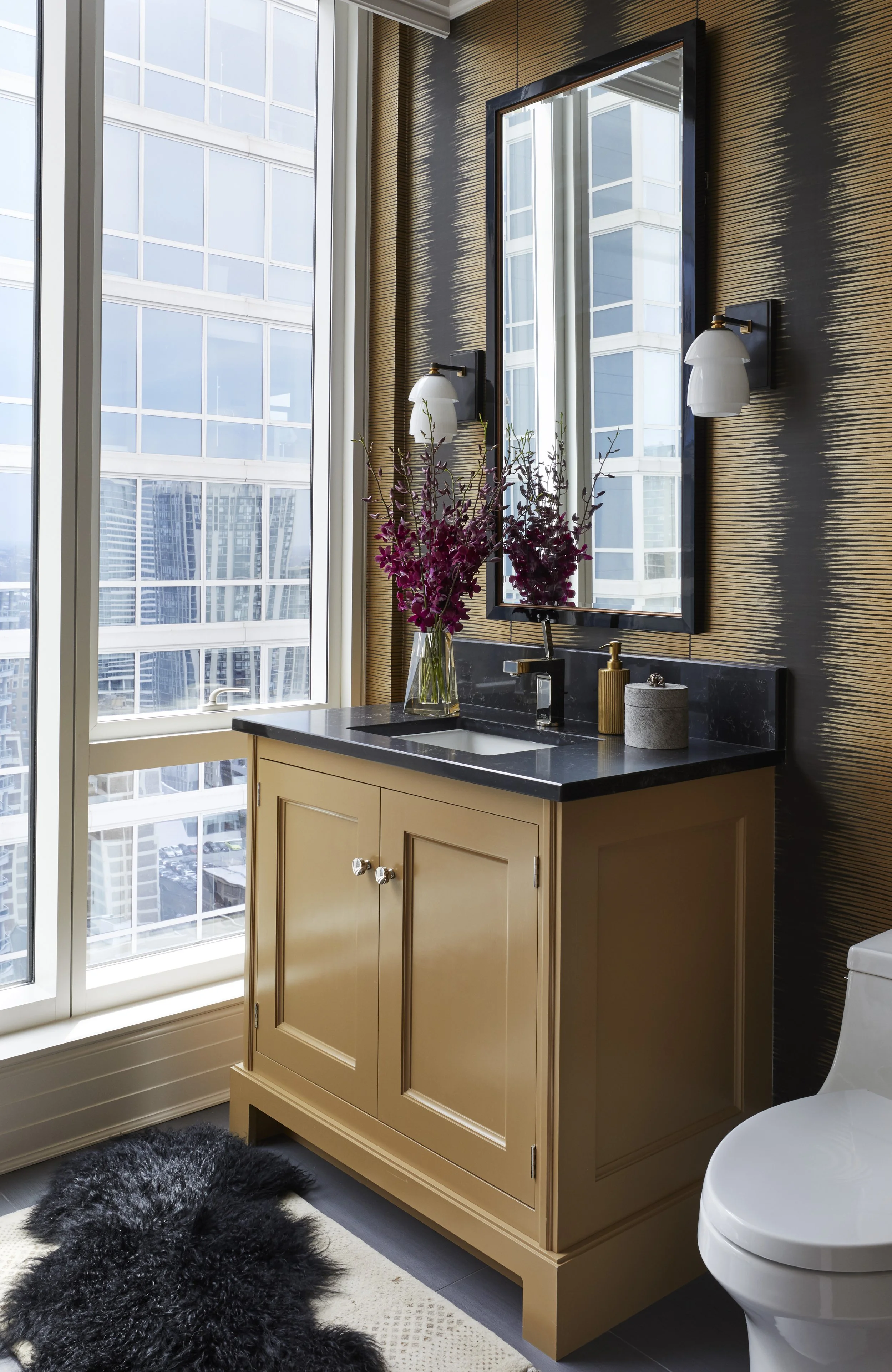 A bathroom with a large window, a vanity with a black countertop, a mirror, and decorative items including a vase with purple flowers, a soap dispenser, and a container. There is a black fluffy rug on the floor and a white toilet next to the vanity. 