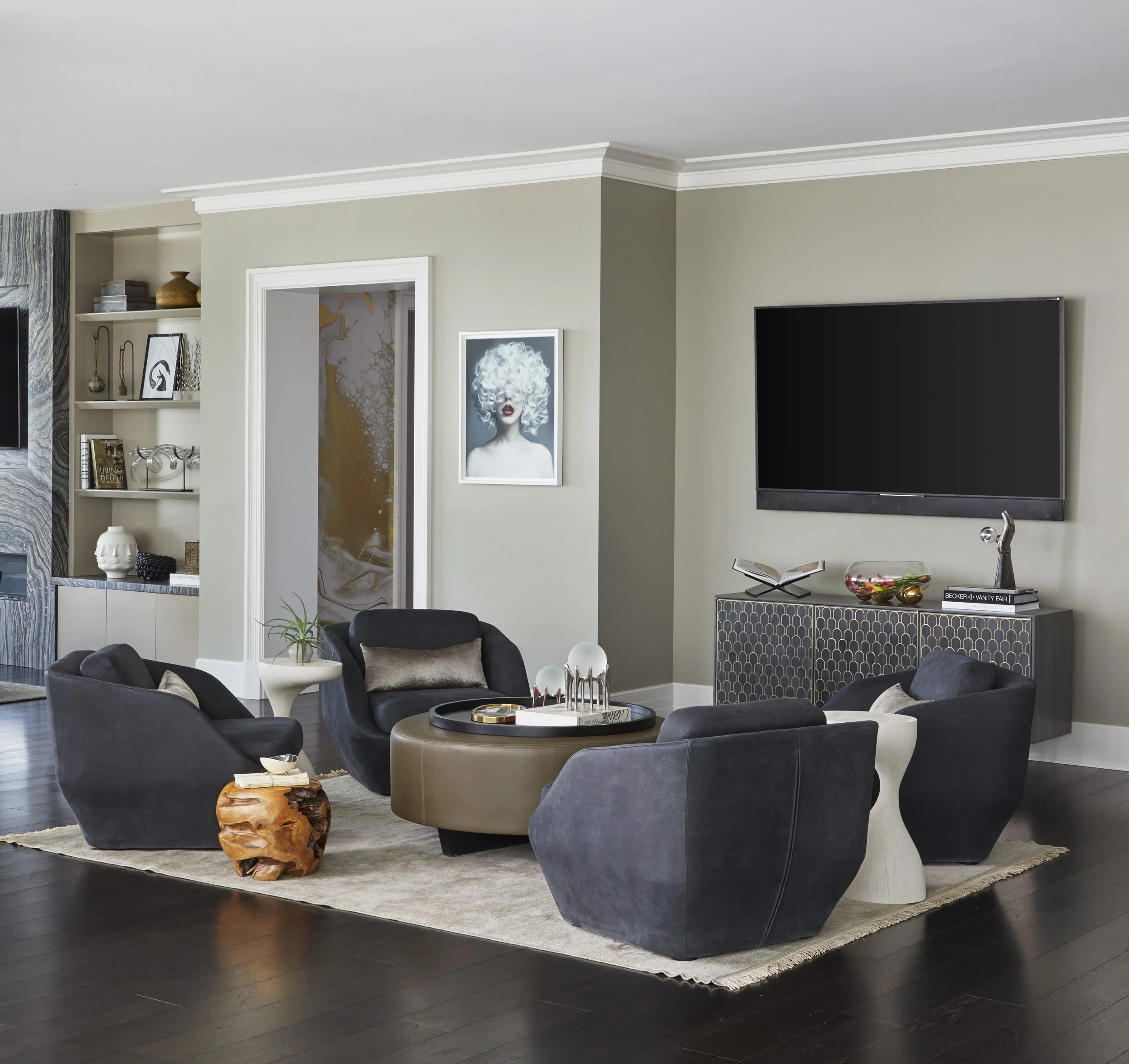 A modern living room with black armchairs surrounding a round coffee table on a beige rug, a white sculpted bust, a side table with a book, and a distinctive wooden side table. Artworks and decorative objects are on the shelves and wall, with a large