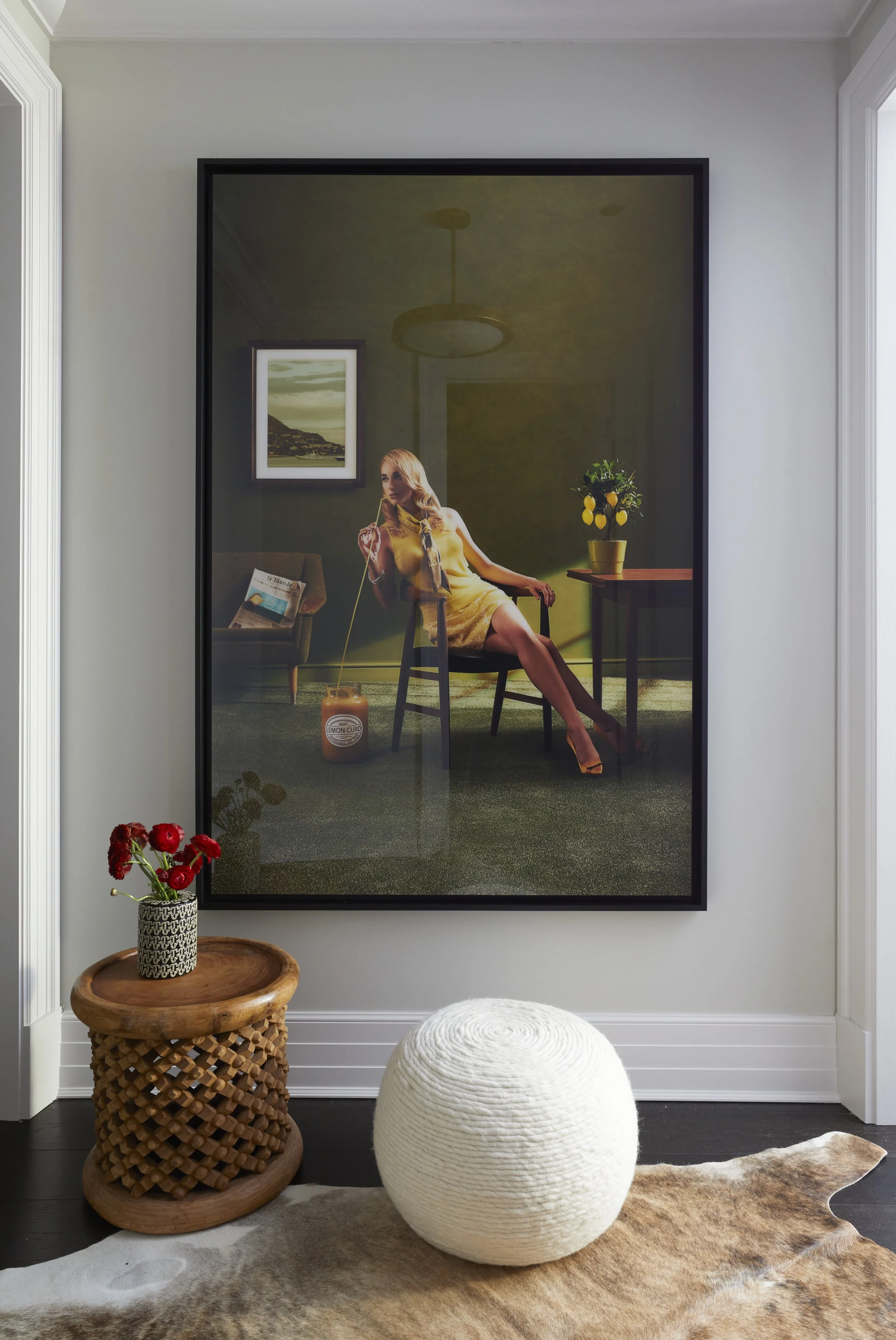 Interior scene with a framed picture on the wall, a woman sitting in a chair holding a drink, a small table with a lemon tree, a side chair with a newspaper, a decorative jar, a cowhide rug, a woven round ottoman, and a vase with red flowers on a woo