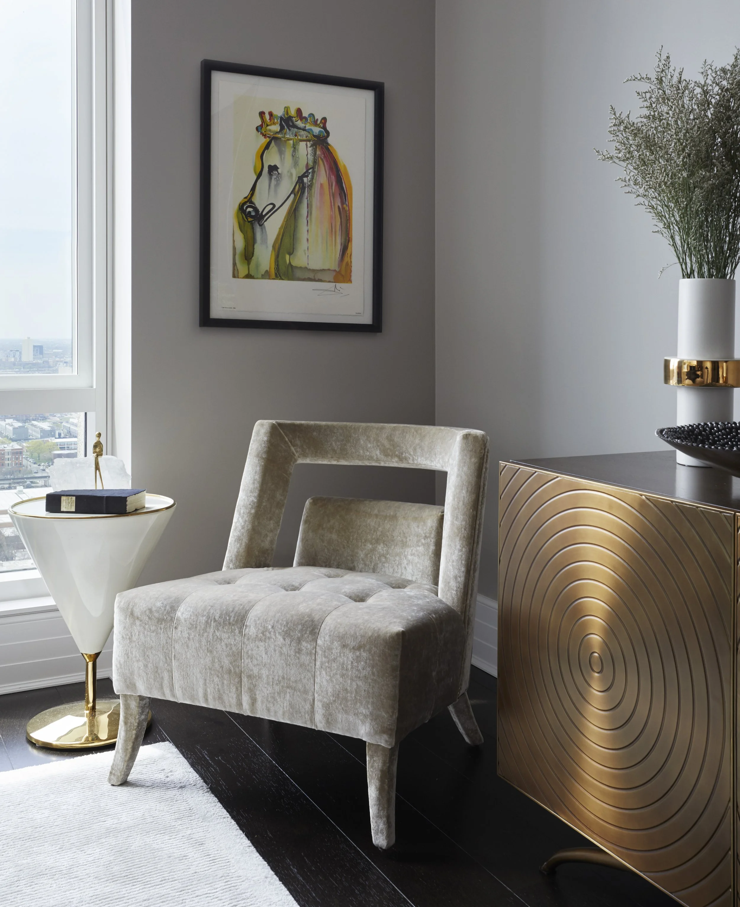 Interior of a modern living room featuring a beige velvet armchair, a white side table with a book and decorative object, large cityscape window, framed abstract horse artwork on the wall, a glossy black and gold sideboard, and a tall white vase with
