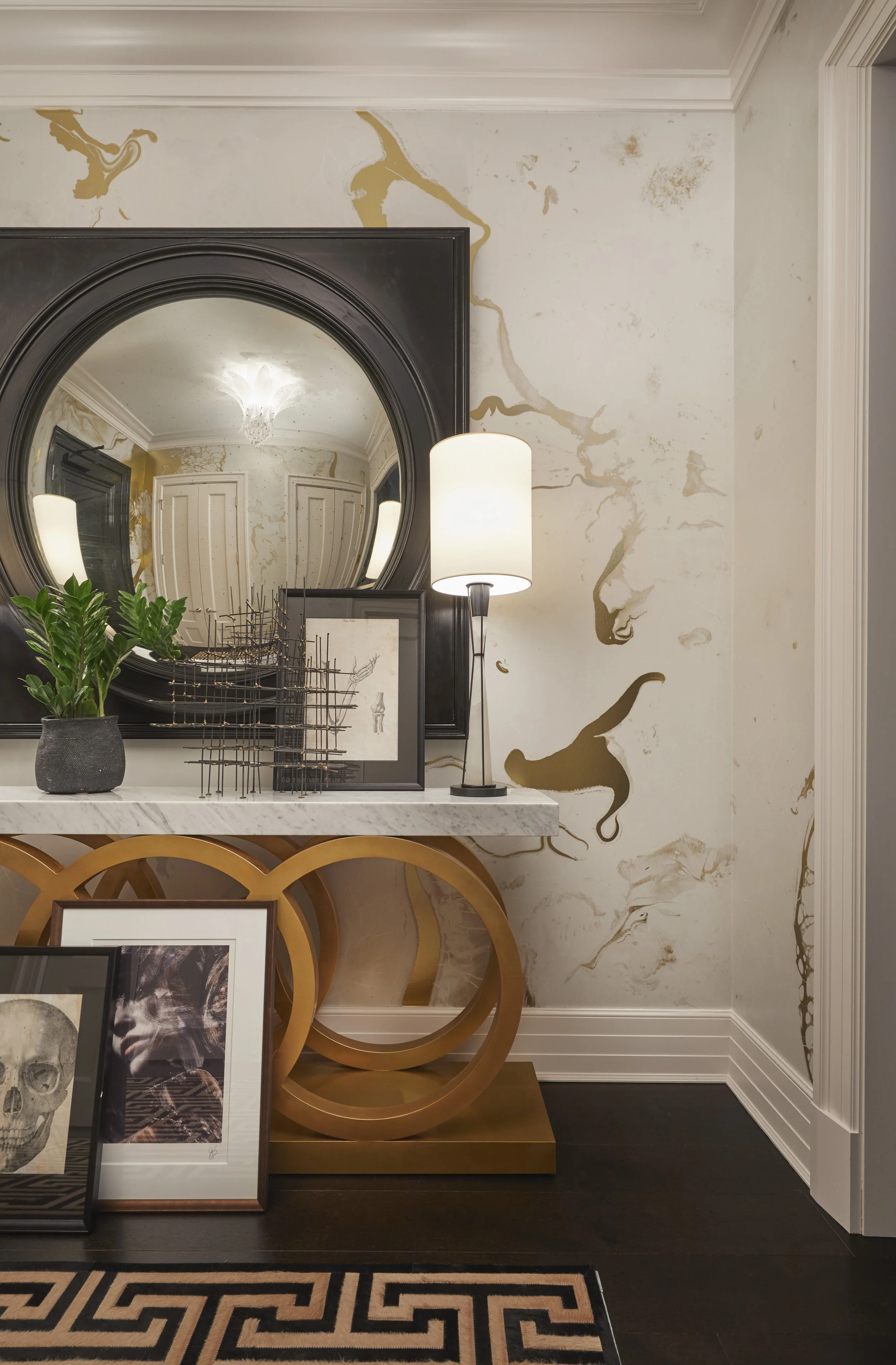 Living room corner with a large mirror, a table with framed artwork, a decorative gold circular table, a floor lamp, and a wall with beige and gold marbled wallpaper.