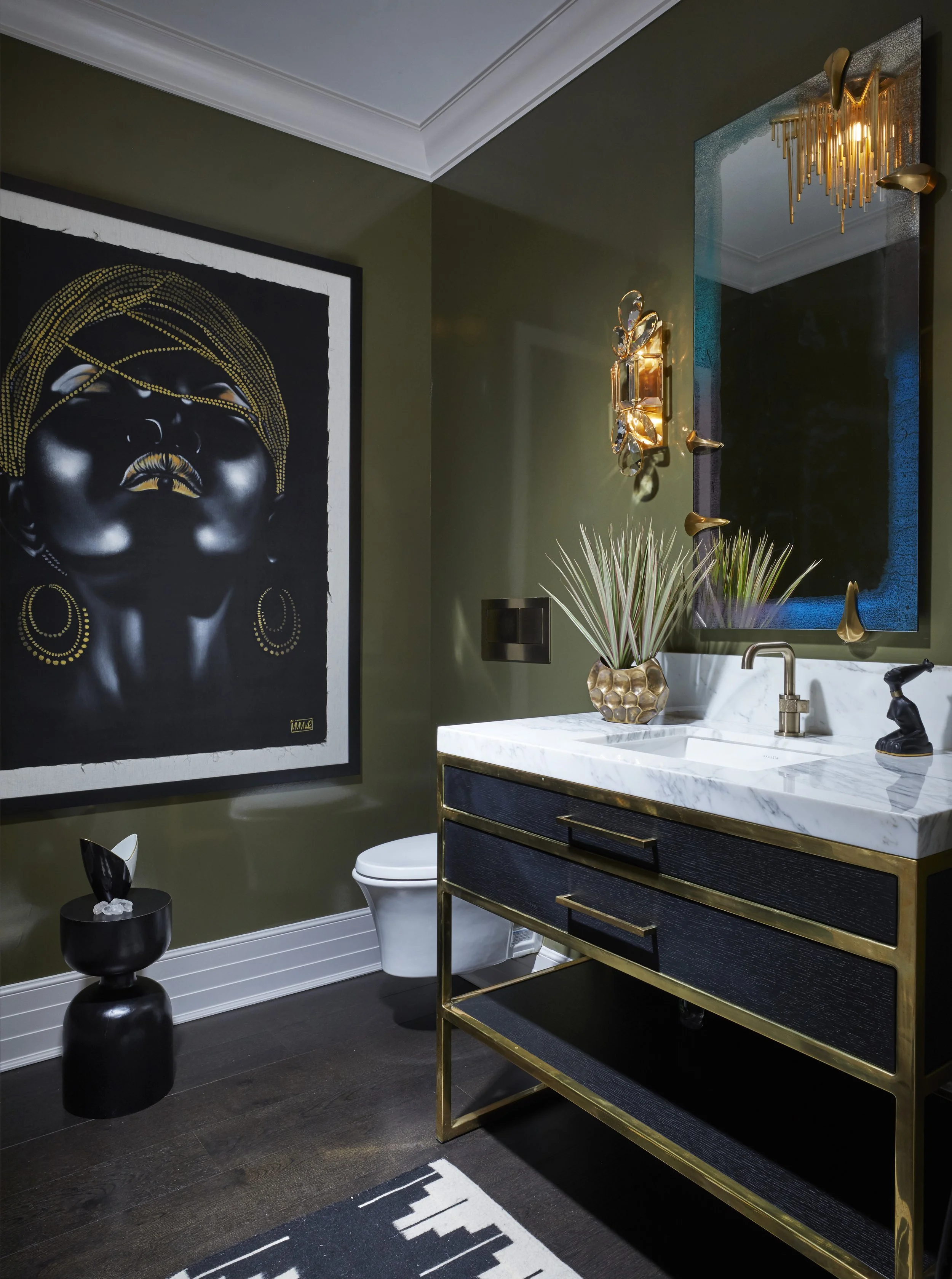 A modern bathroom with dark wood floors, olive green walls, and white crown molding. Features a black and white abstract art piece on the wall, a marble countertop with a sleek faucet, a decorative plant in a geometric gold vase, and gold accent ligh