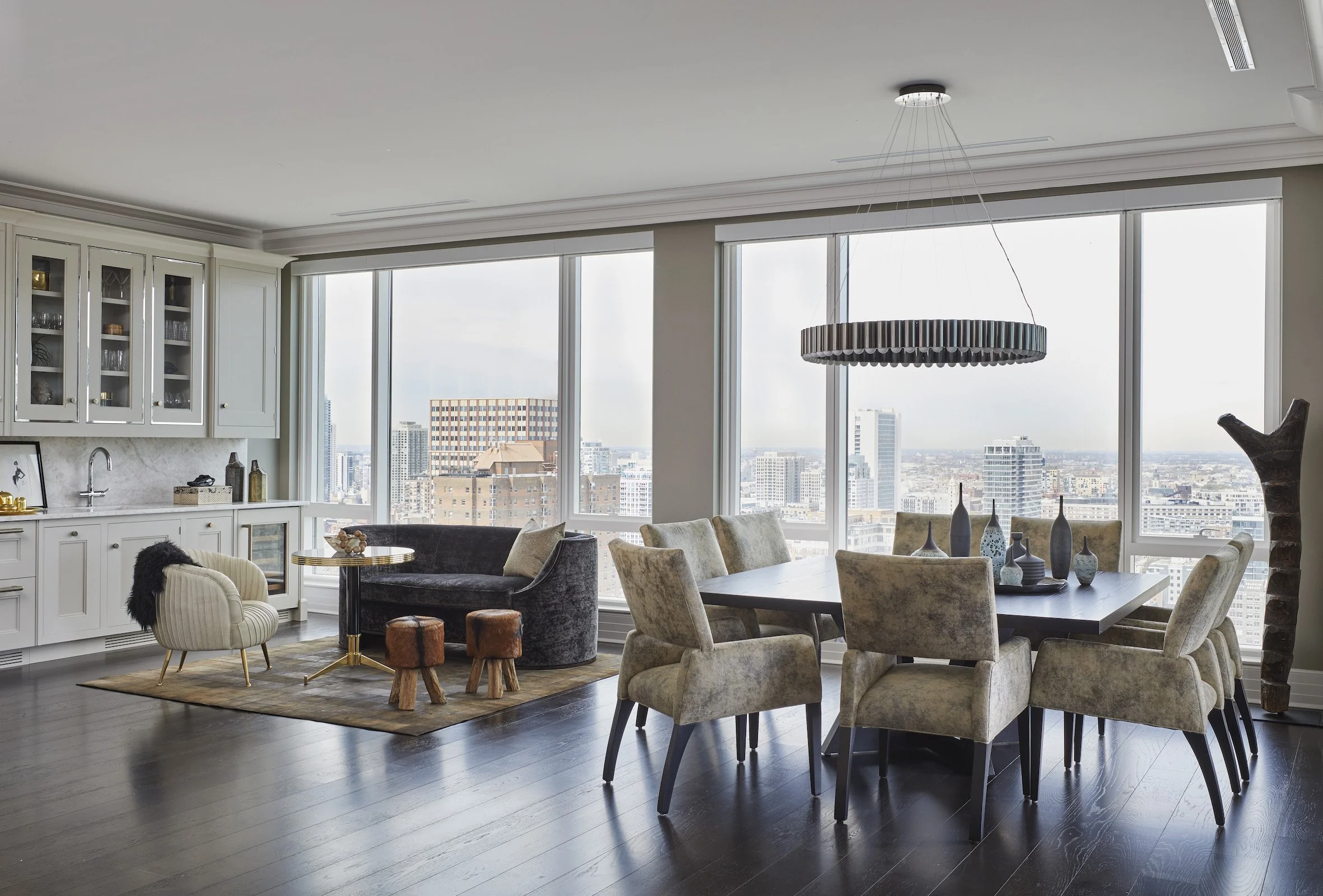 Modern high-rise apartment living room with large windows showing cityscape, featuring a dining table with beige upholstered chairs, a black velvet armchair, a white cabinet with glass doors, decorative vases, and a contemporary chandelier.