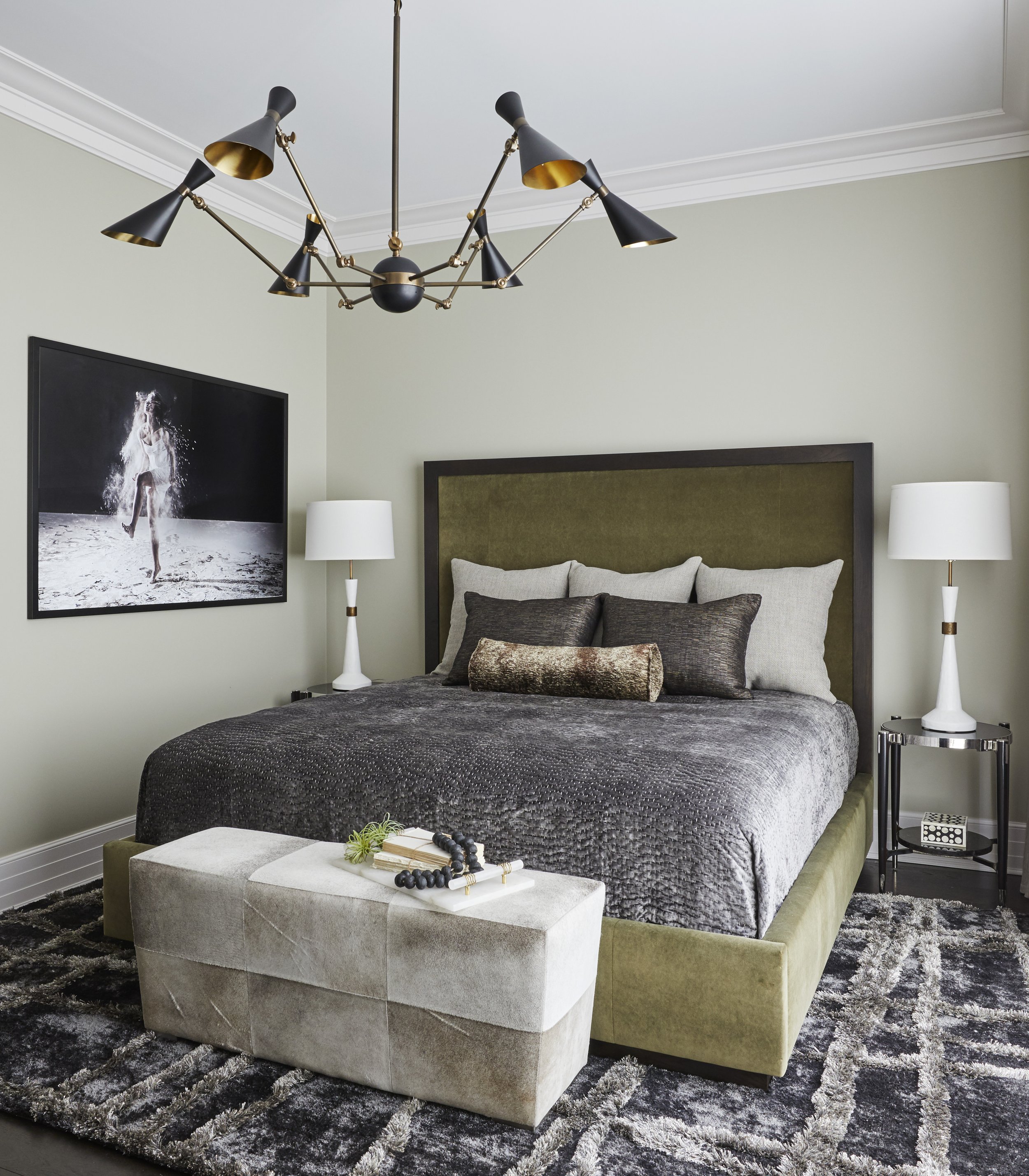 Modern bedroom with a green upholstered bed frame, gray bedding, black and gray pillows, a white nightstand, and lamps. A black framed artwork of a dancing woman and a black chandelier hang above the bed. A white tufted bench with decor and a black a