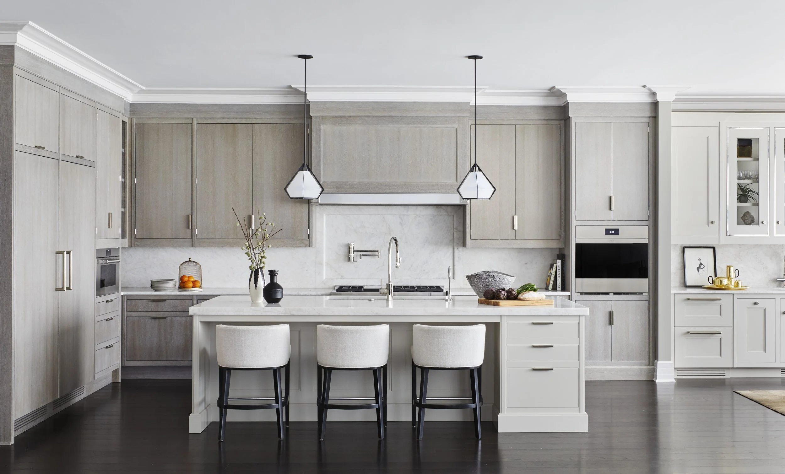 Modern kitchen with light wood cabinetry, white countertops, and a central island with three chairs. Two pendant lights hang above the island. The kitchen has a built-in oven, microwave, and a small open shelf with decorative items. A vase with flowe