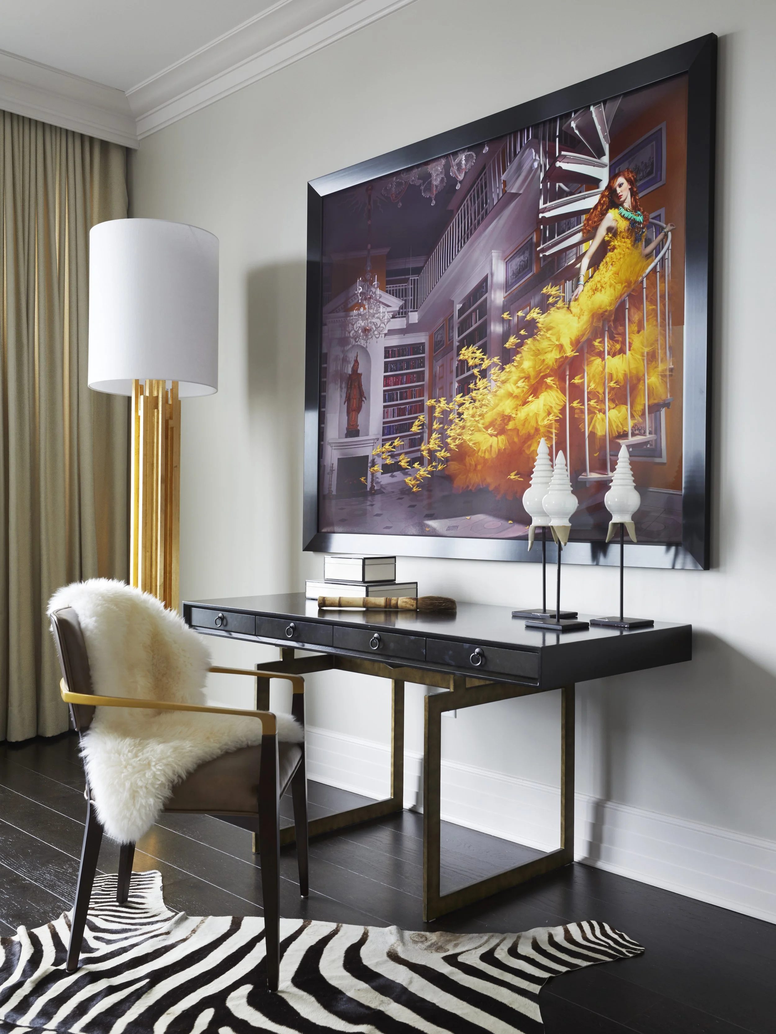 A modern home office space featuring a black desk, a chair with a sheepskin cover, a zebra-print rug, a large framed artwork of a woman in an orange dress on the wall, white decorative objects, and a lamp with a gold base.