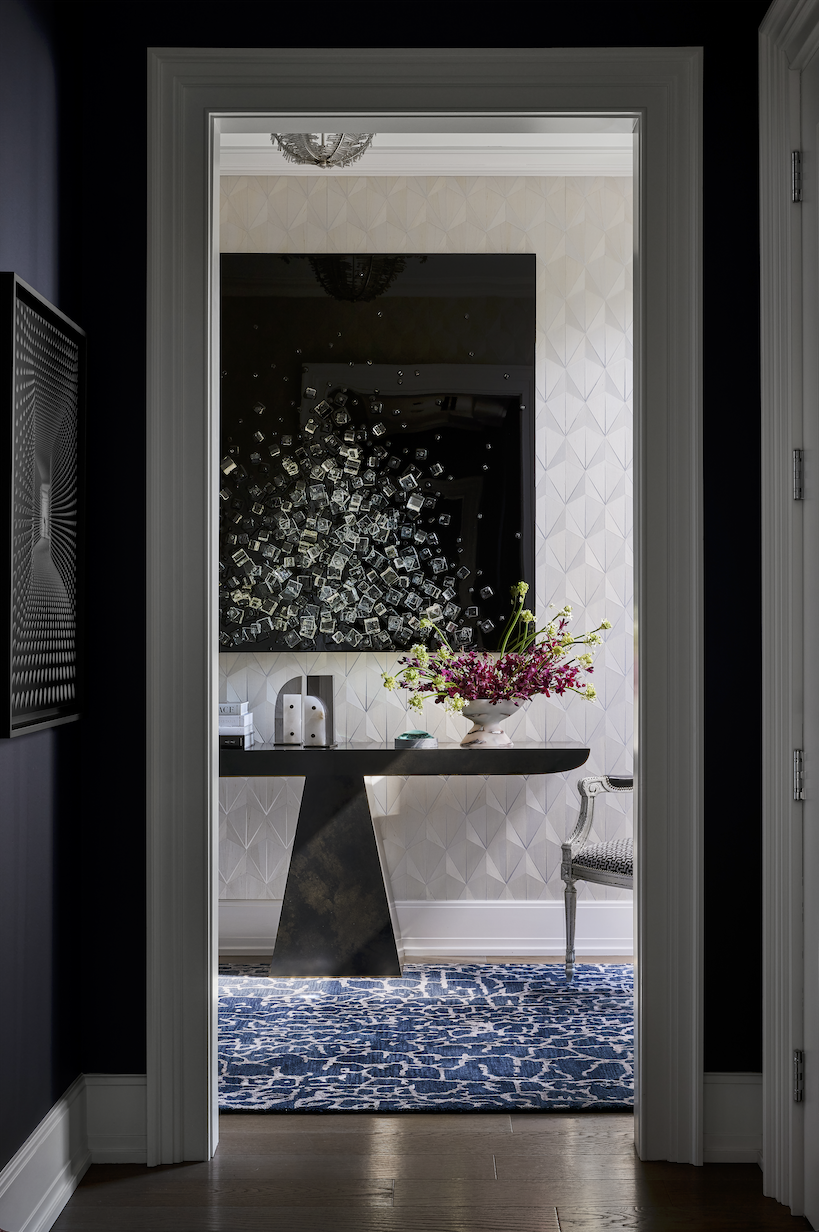 View through a doorway into a room with a black table, a large floral arrangement in a white vase, a patterned armchair, a wall-mounted television, and a chandelier reflected in the ceiling mirror. The room features patterned wallpaper and a blue and