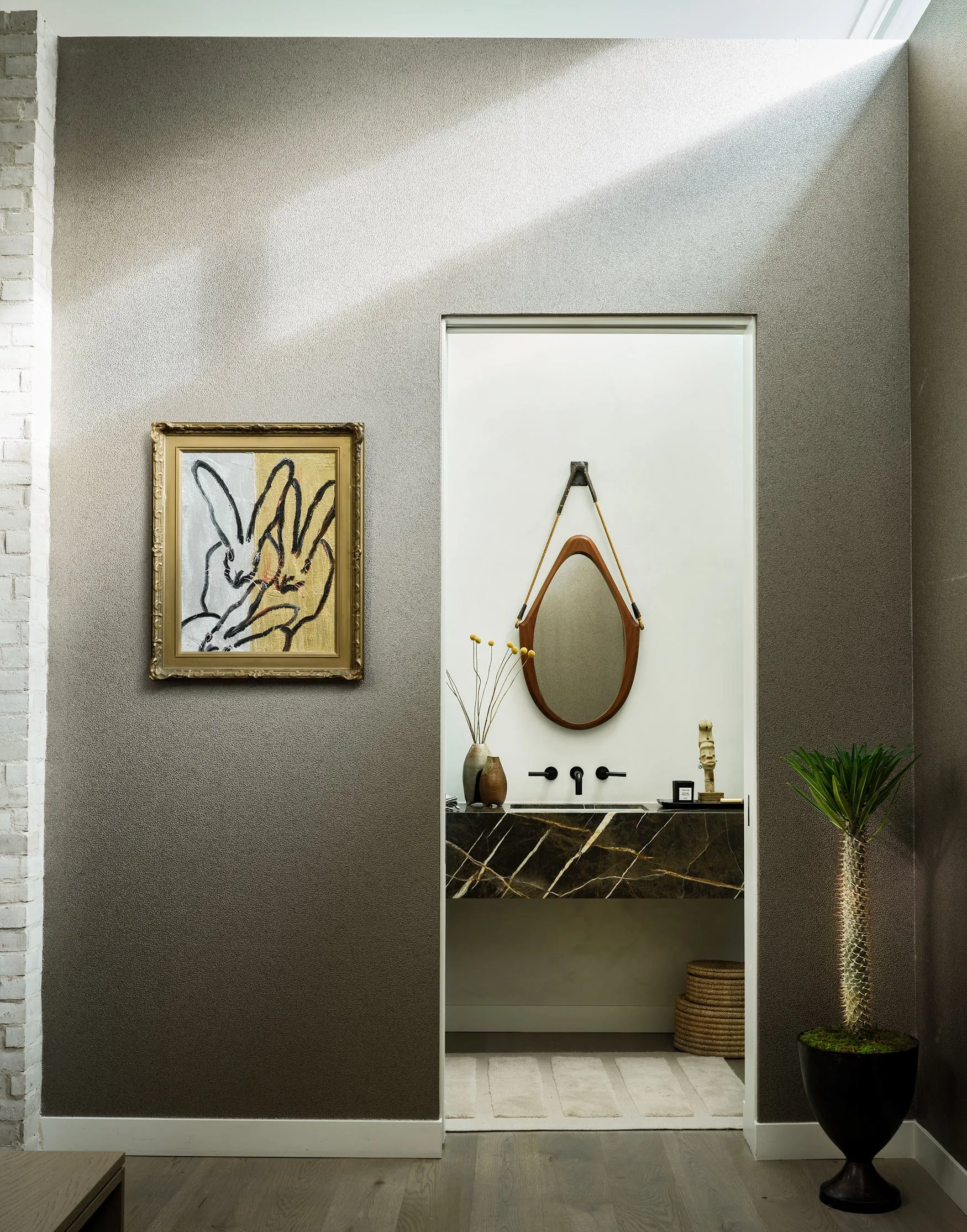 Interior view of a modern bathroom with a white door, a mirror with a wooden frame, and decorative items including vases and a plant in a black pot.