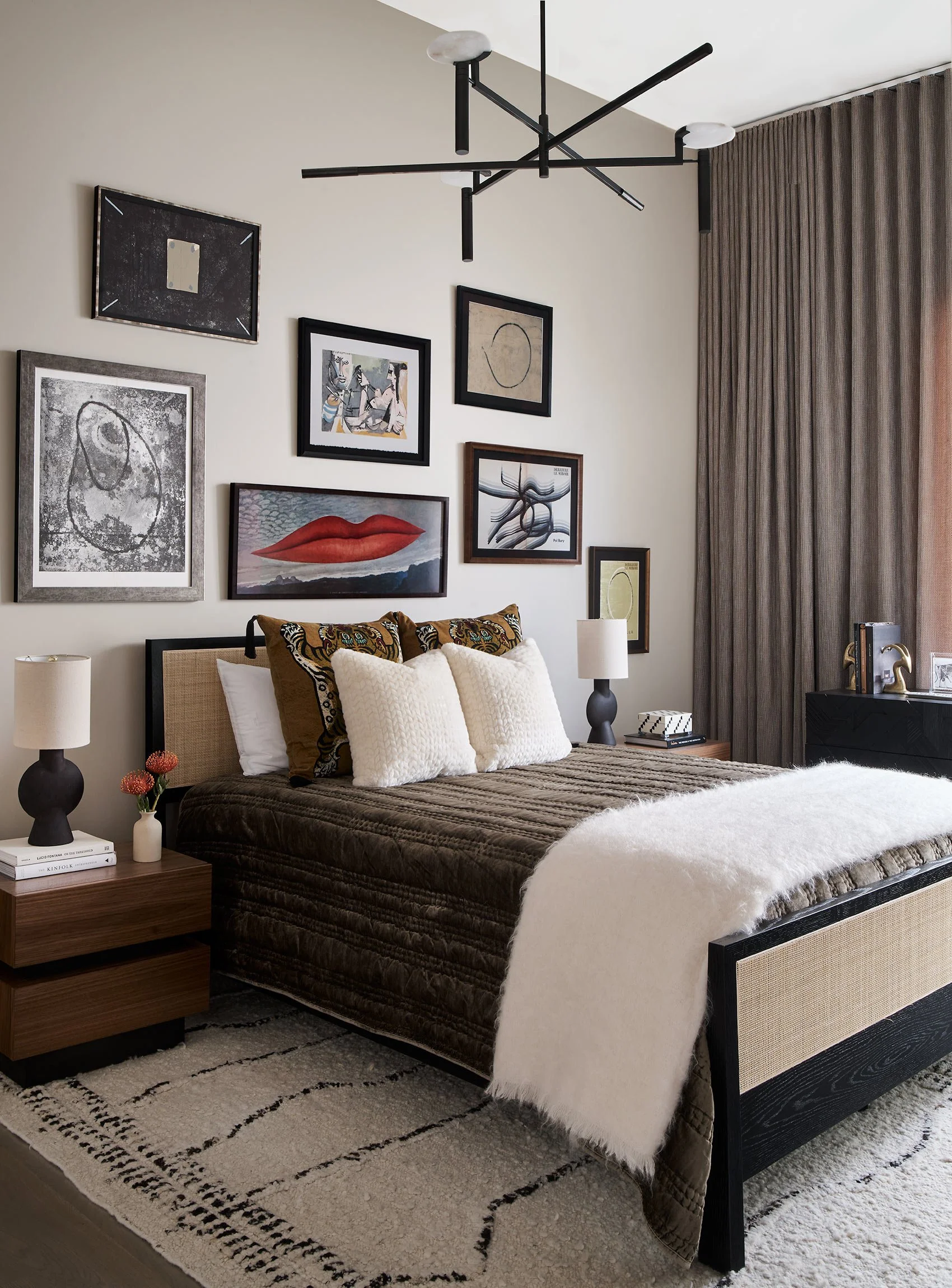 A modern bedroom with a black and beige bed, white pillows, and a white throw blanket. Wall art includes abstract and figurative pieces. A black bedside lamp, a wooden nightstand with books and a small vase, and a patterned area rug complete the deco