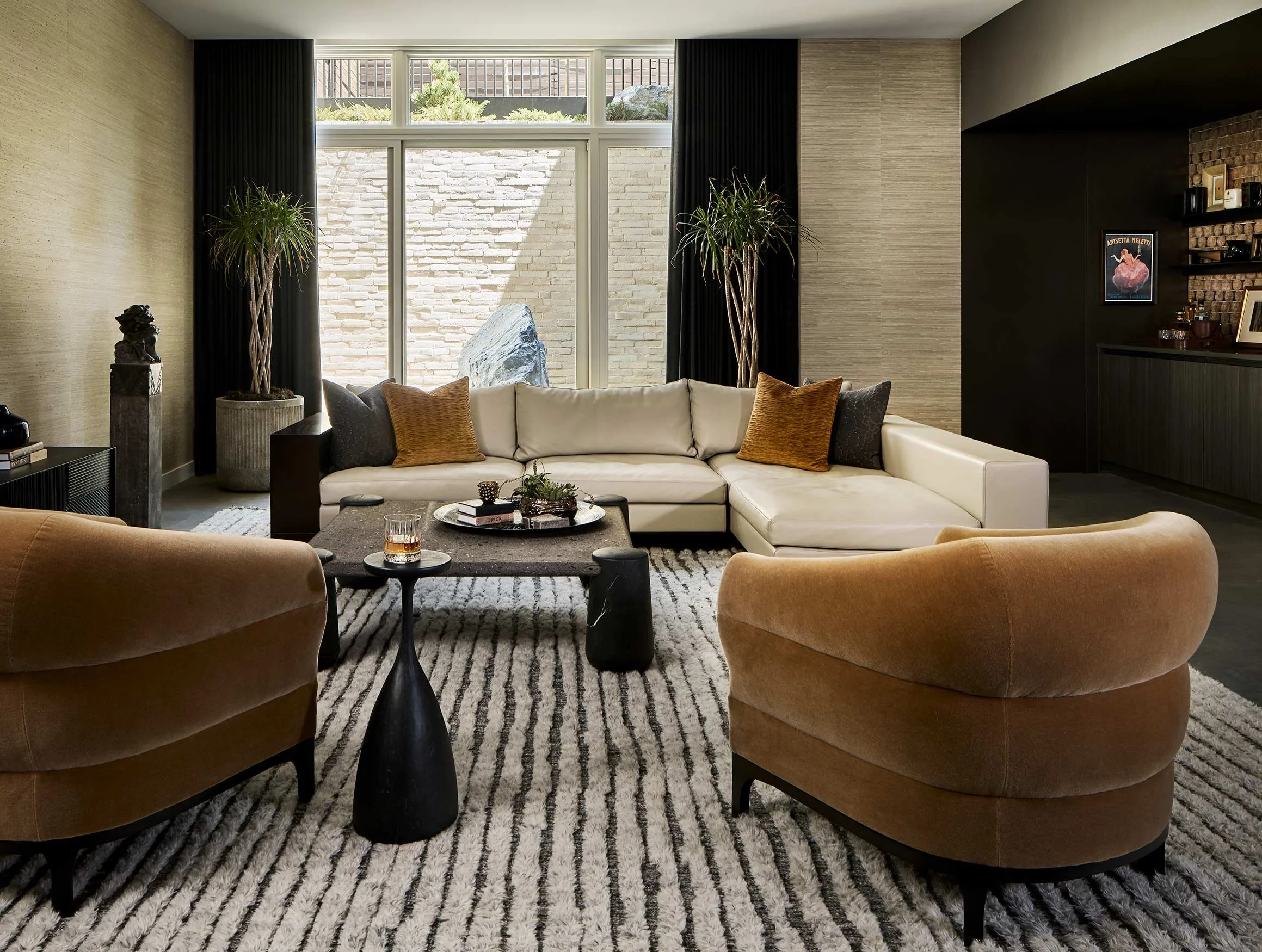 Modern living room with beige sectional sofa, two brown armchairs, black coffee table, black and beige rug, large window with black curtains, two tall indoor plants, brick wall with art and bar area.