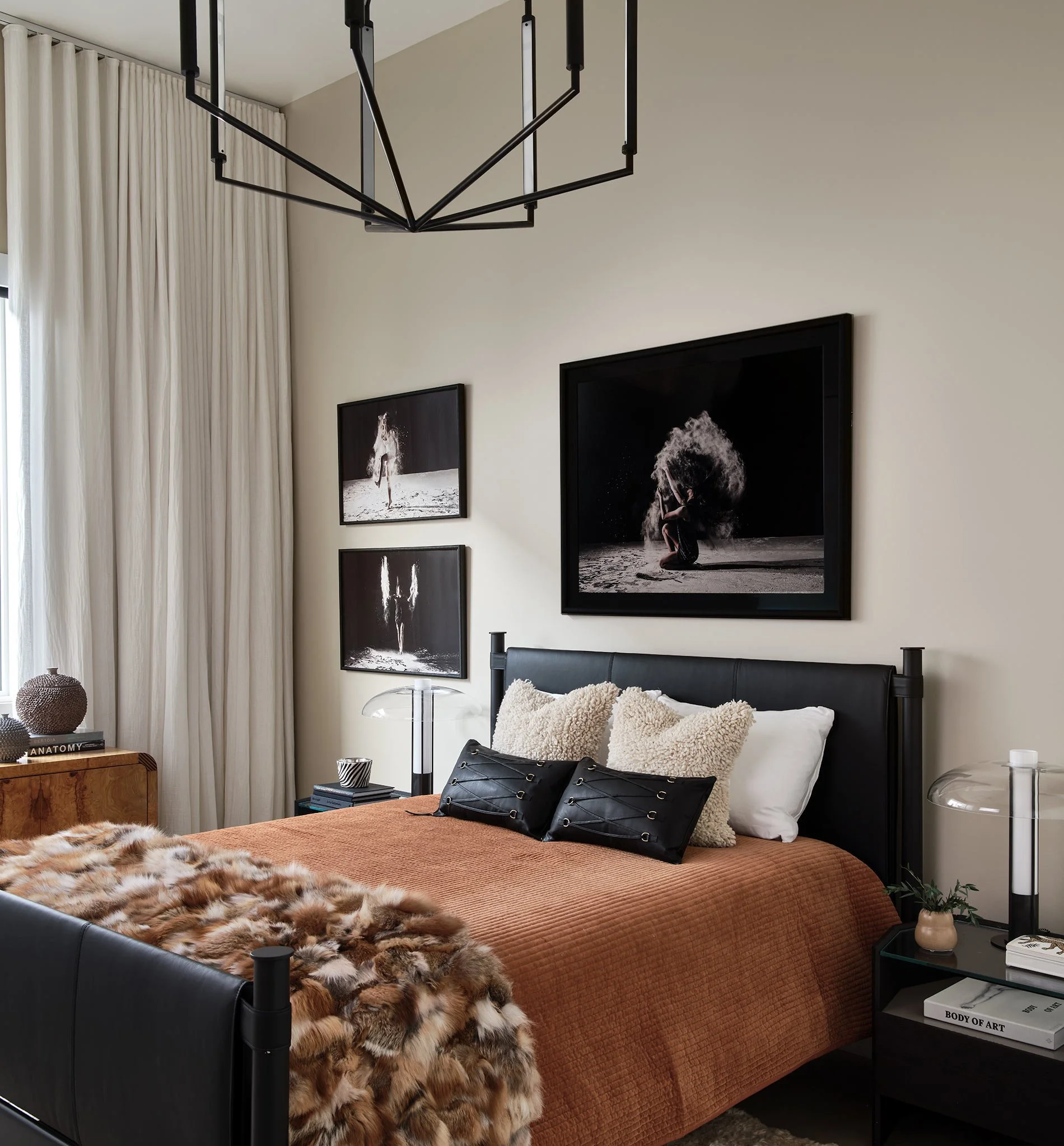 A bedroom with a black bed frame, orange bedspread, and furry throw blanket. Black pillows with straps, small side table, and framed black-and-white artwork of a person on sand. Off-white curtains and minimal decor.