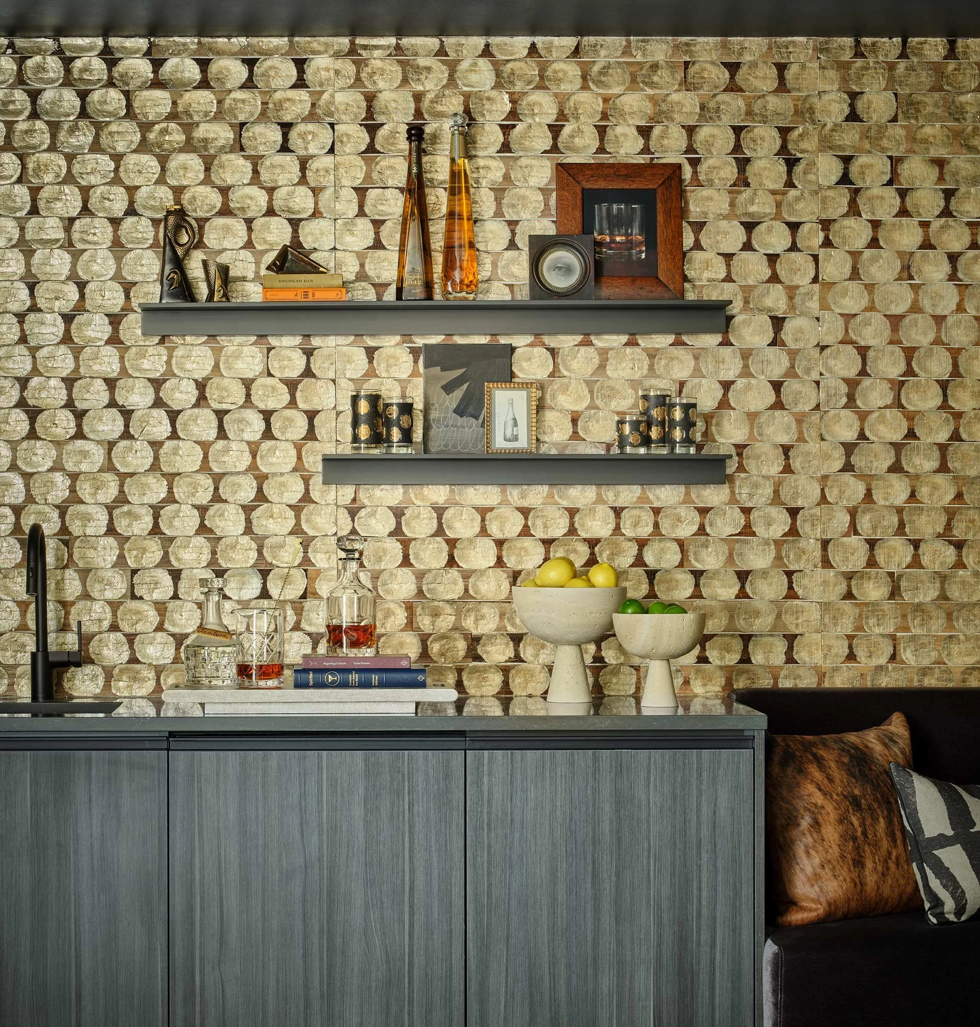 A kitchen with a backsplash made of round, beige stone tiles. Two gray floating shelves hold decorative items, including glass bottles, books, and framed photos. The lower shelf has black and gold decorative cups, and a bowl of lemons, with part of a
