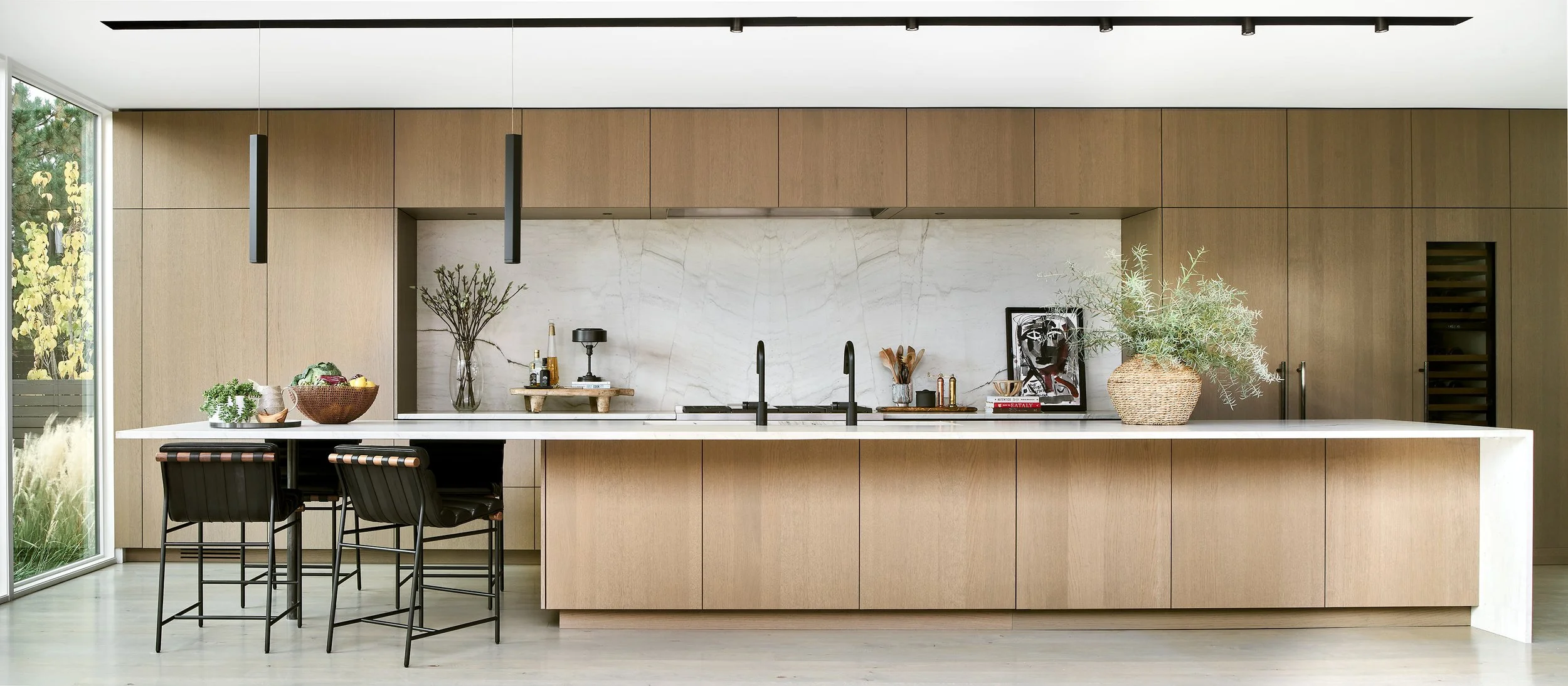 Modern kitchen with wooden cabinets, white marble backsplash and island, black fixtures, decorative plants, and stools.