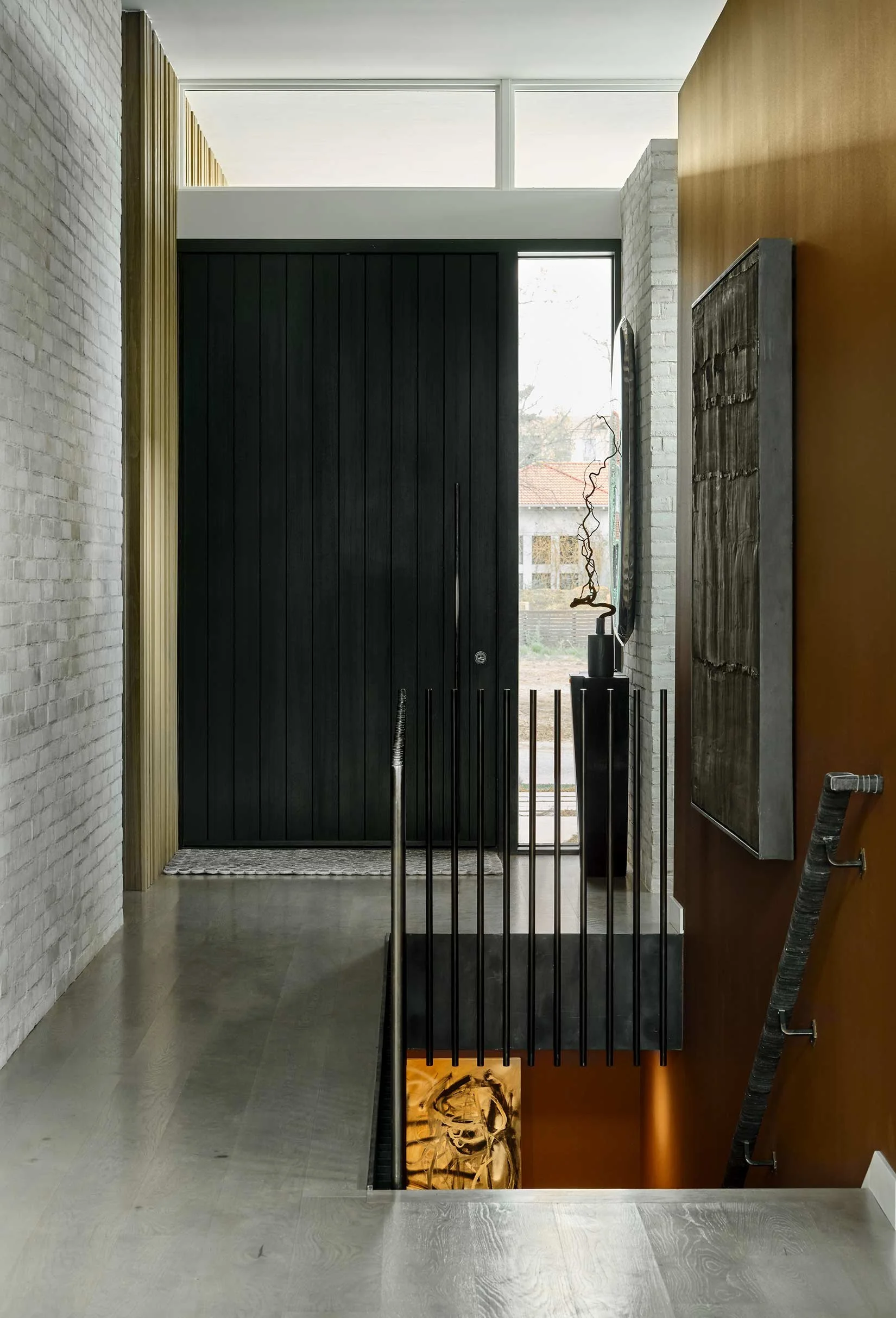 Modern entryway with black door, white brick wall on the left, wooden wall on the right, decorative objects including a tall black vase with a curly dried branch, and stairs leading downward with a painting at the bottom.