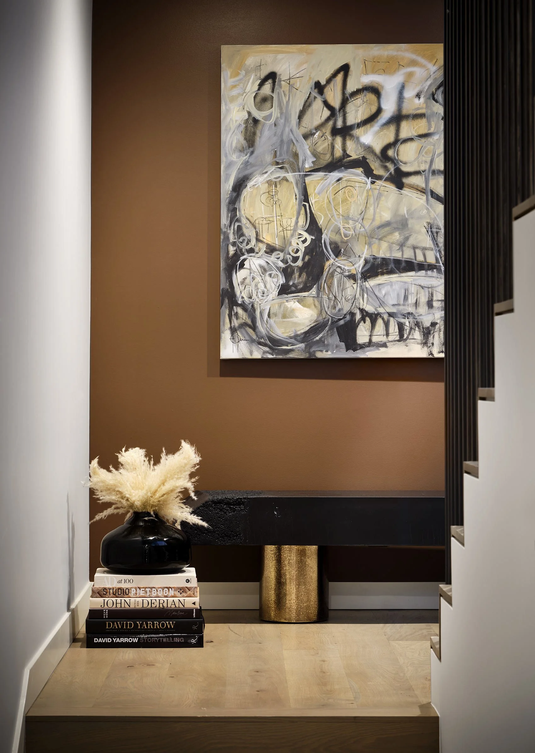 An interior space with a modern decor, featuring a stack of books with titles about art and storytelling on a small white table, a black vase with beige pampas grass, a large abstract painting on the wall, a gold textured cylindrical side table, and 