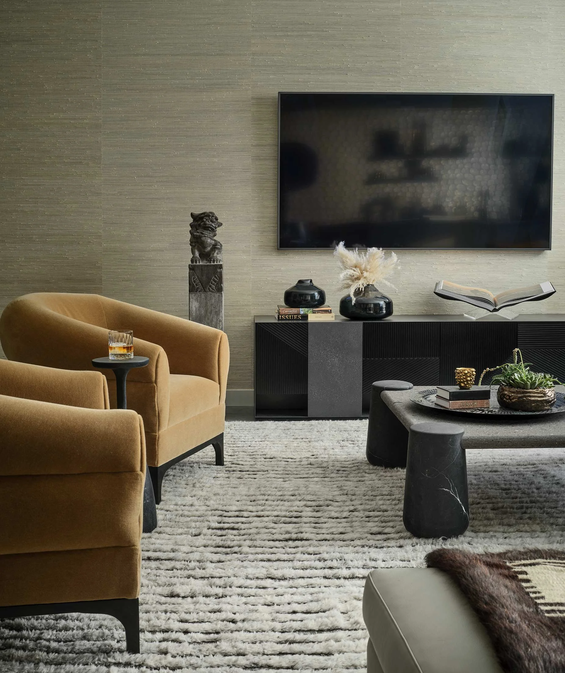 Modern living room with beige sofa, textured beige wall, wall-mounted flat-screen TV, black decorative pieces, a sculpture of a lion on a pedestal, books, and a coffee table with a plant and a candle.