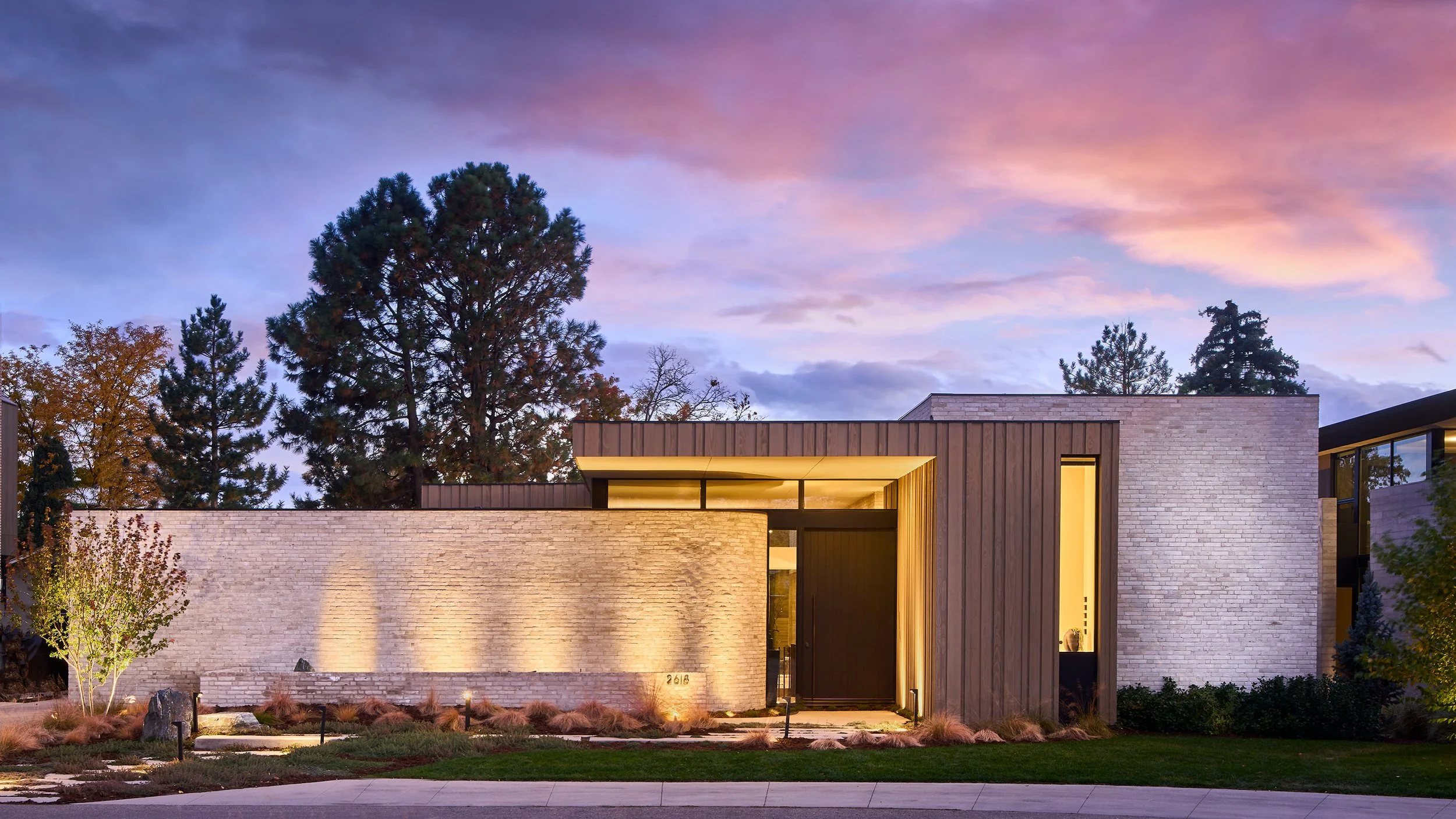 Modern house with a flat roof and large windows, illuminated from inside, surrounded by trees and landscaped yard, during dusk with a colorful sky.