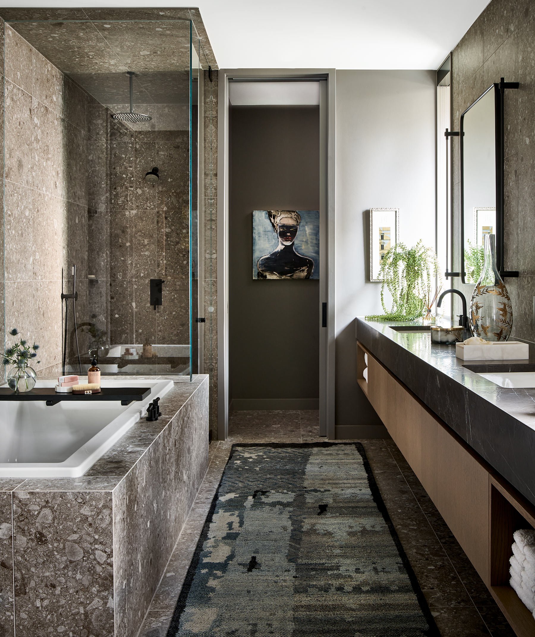 Modern bathroom with a walk-in shower featuring stone tiles, a bathtub with toiletries and a black tray, a tall mirror, a wooden vanity with a black countertop, decorative vases, a potted plant, and a patterned rug.