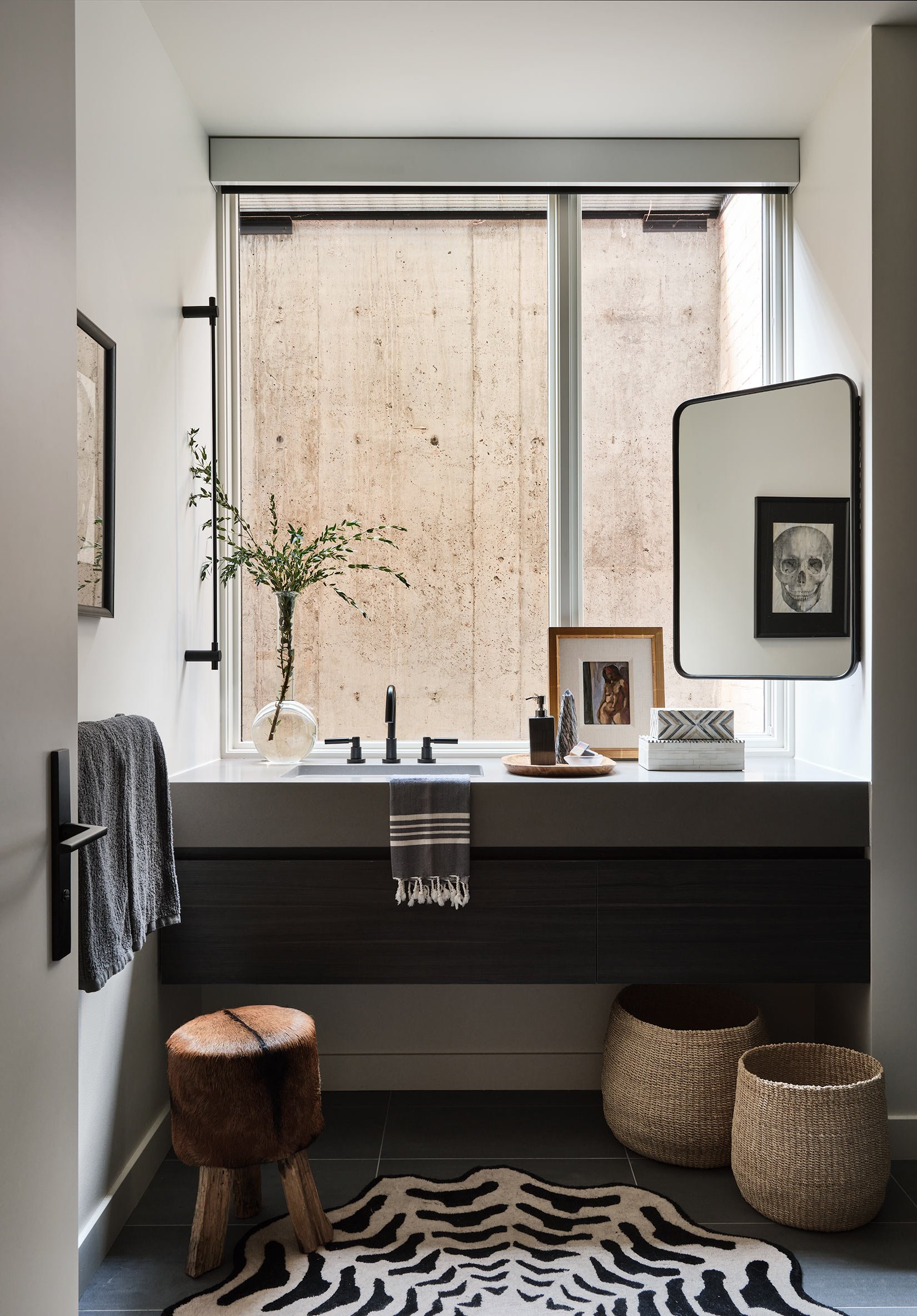Modern bathroom vanity with a black cabinet, a white countertop, and a black faucet. Decor includes framed artwork, a glass vase with green branches, and various small objects. The space has a large window with a concrete wall background, zebra-patte