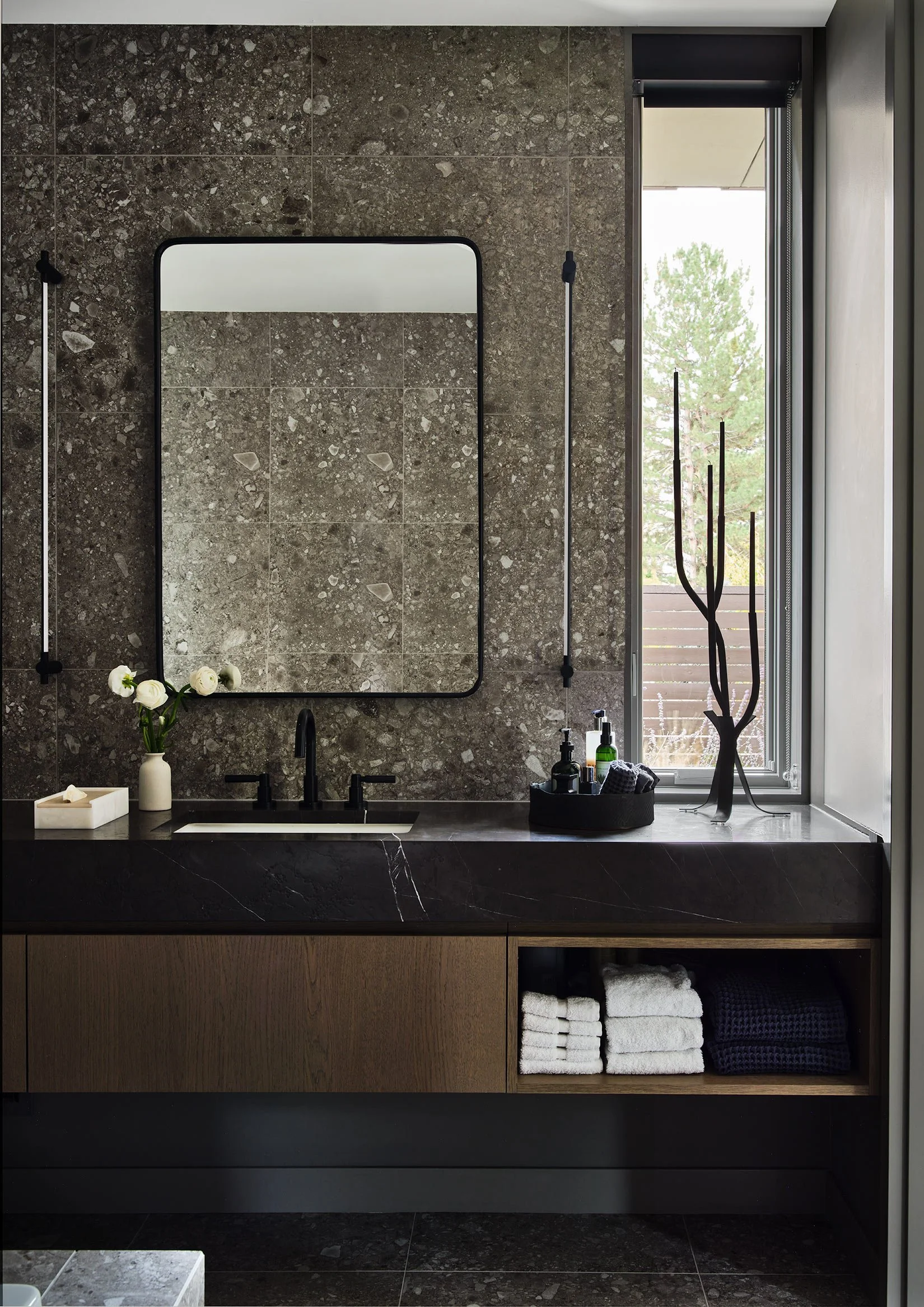 Modern bathroom vanity with a dark marble countertop, a white vase with flowers, a black faucet, and neatly folded white and dark towels on an open shelf. The background features a large mirror, textured tile wall, and a window with a view of trees o