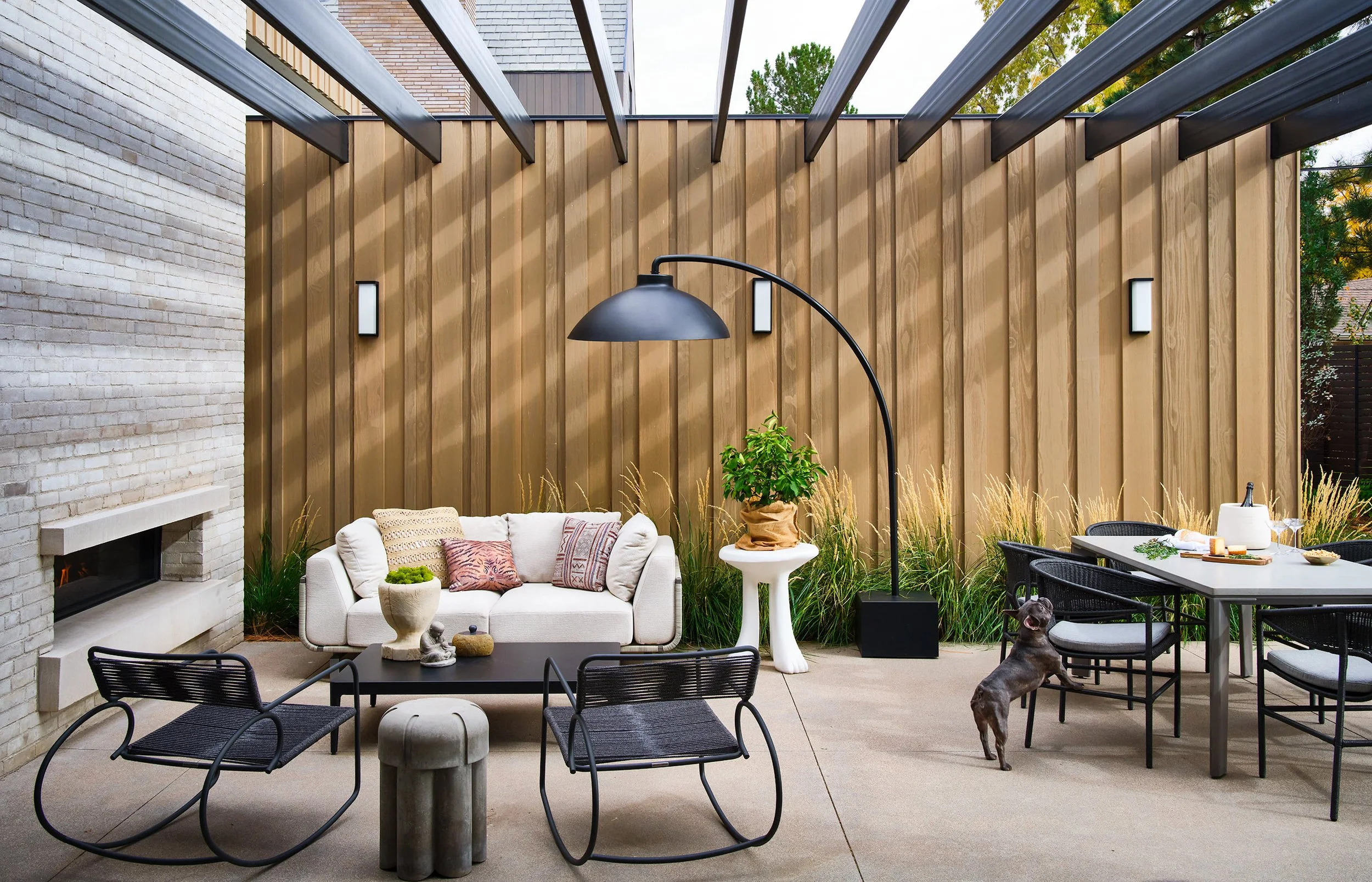 Outdoor patio with modern furniture, including a white sofa with pillows, black chairs, and a table with decorative items. A large black lamp stands near the sofa, and there is a wooden privacy fence with greenery in the background. The patio has a s
