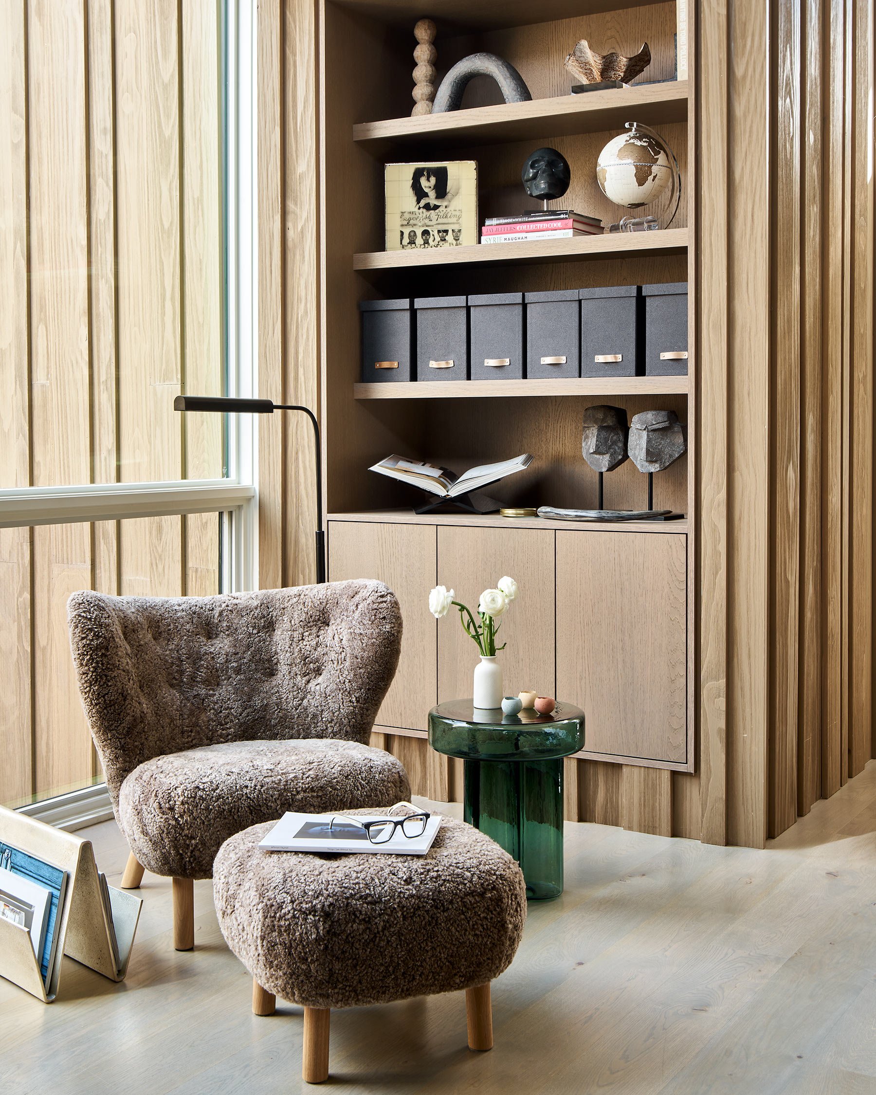 Modern interior living space with a plush, beige armchair, a small round green table with a white vase and decorative objects, and a built-in wood wall unit with decorative objects and books. Natural light through a large window on the left.