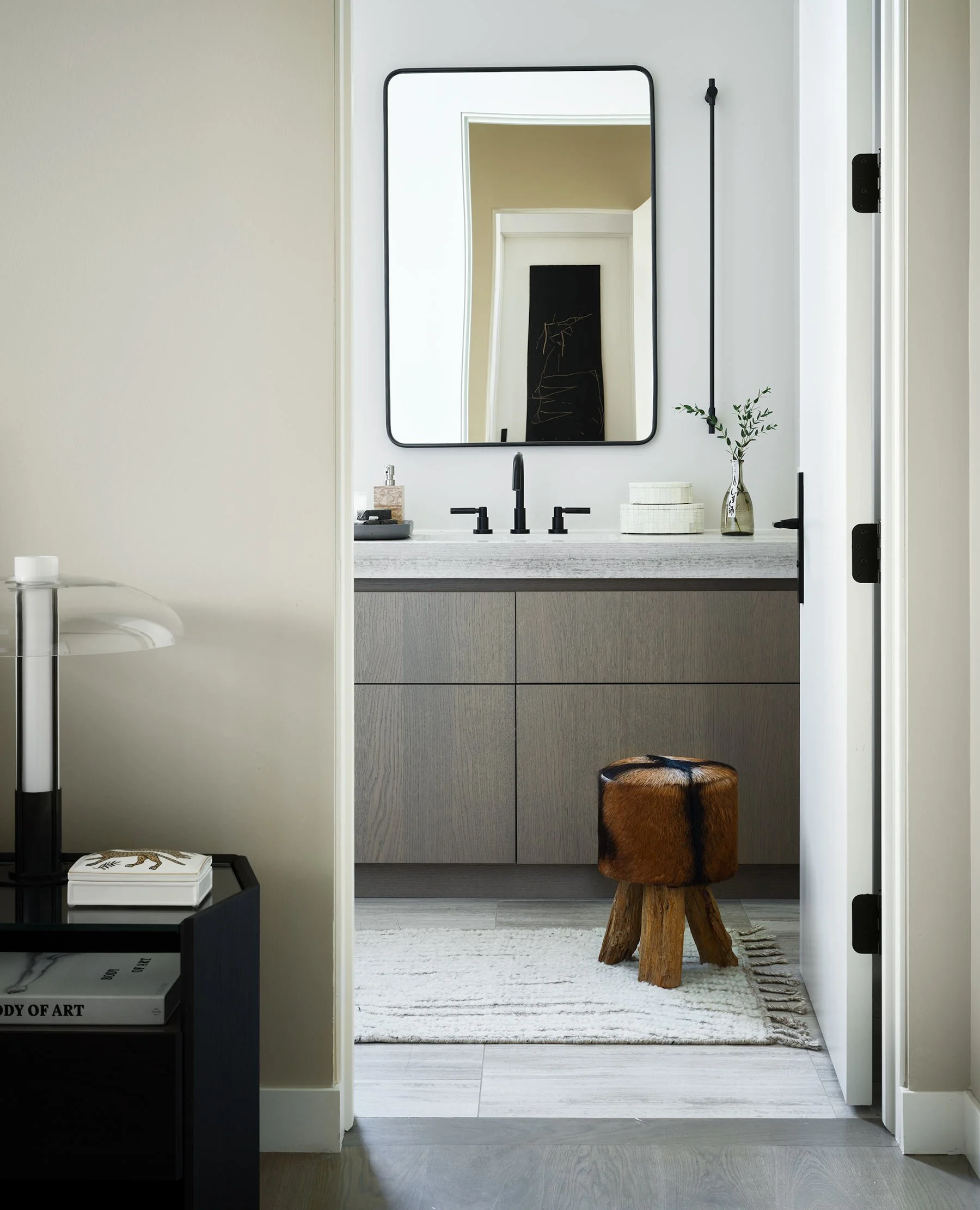 Modern bathroom with a large mirror, black fixtures, and a wooden stool. Decor includes a small plant and woven containers, with a rug on light-colored flooring.