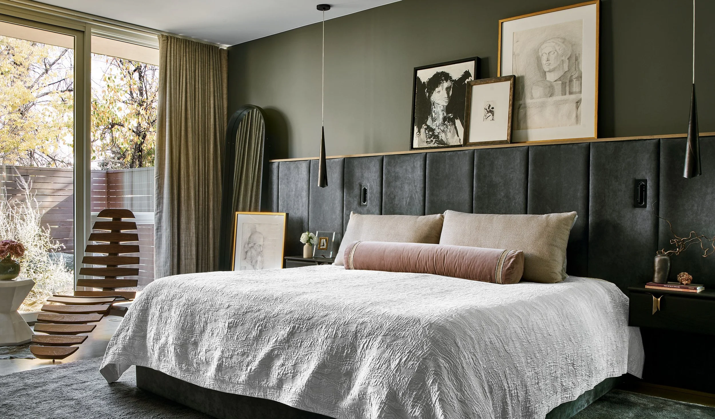 A bedroom with a bed covered in white textured bedding, a beige headboard with a pink bolster pillow, and a small bedside table with decorative items. Artworks hang on a green wall above the bed, and a large window with beige curtains shows trees out