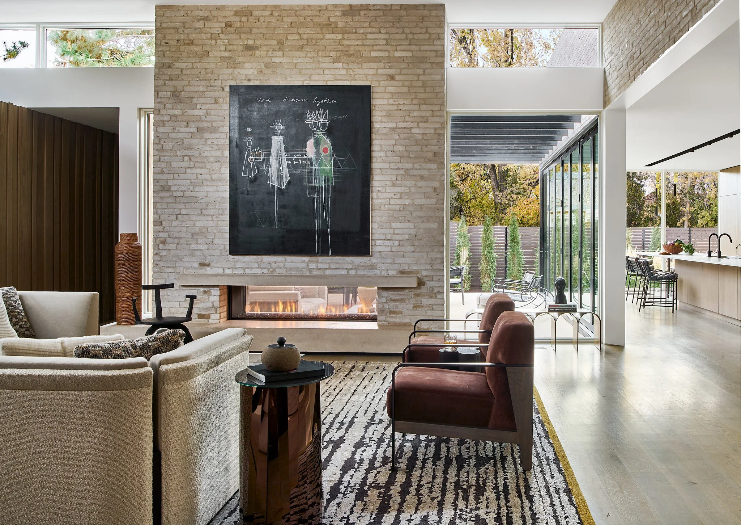 Modern living room with beige sofa, patterned rug, and leather chairs in front of a brick fireplace, large windows showing trees outside, and a view of the kitchen area in the background.