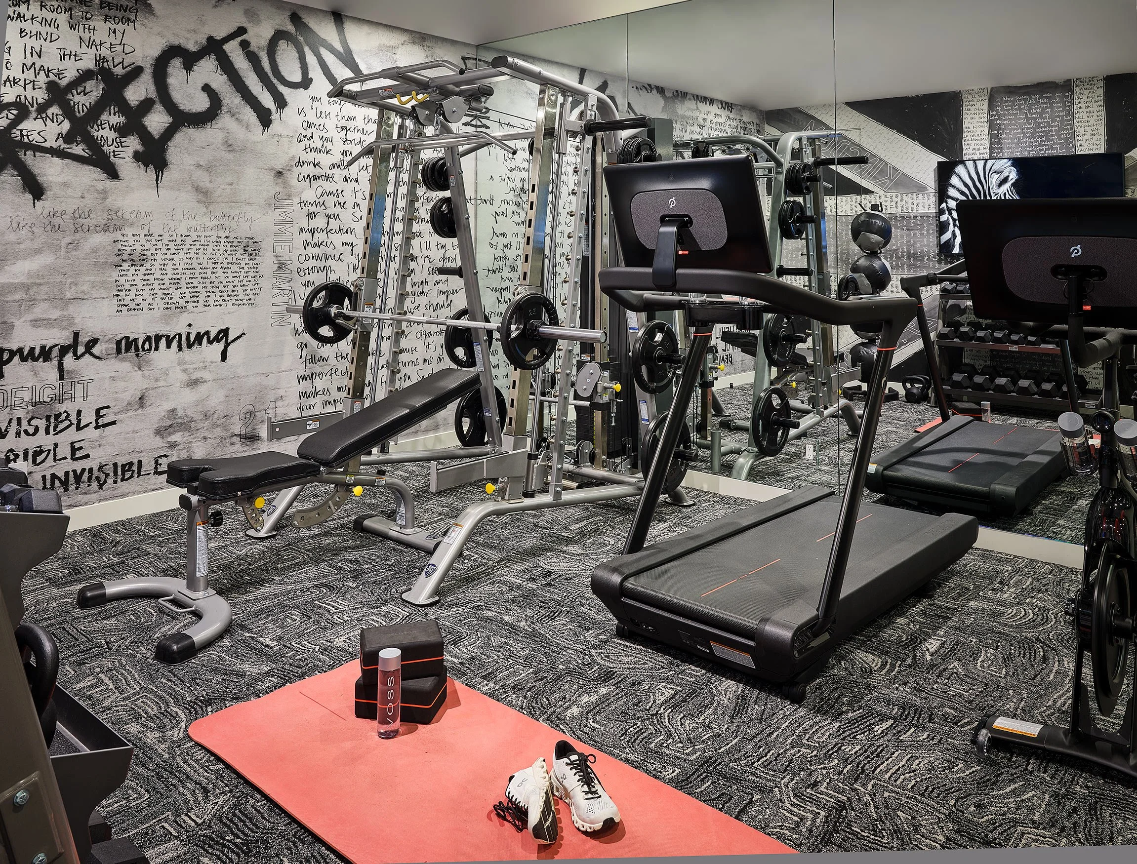 Fitness gym with workout equipment including a treadmill, weight rack, and exercise bench, with a black and white graffiti-style wall and a large mirror.