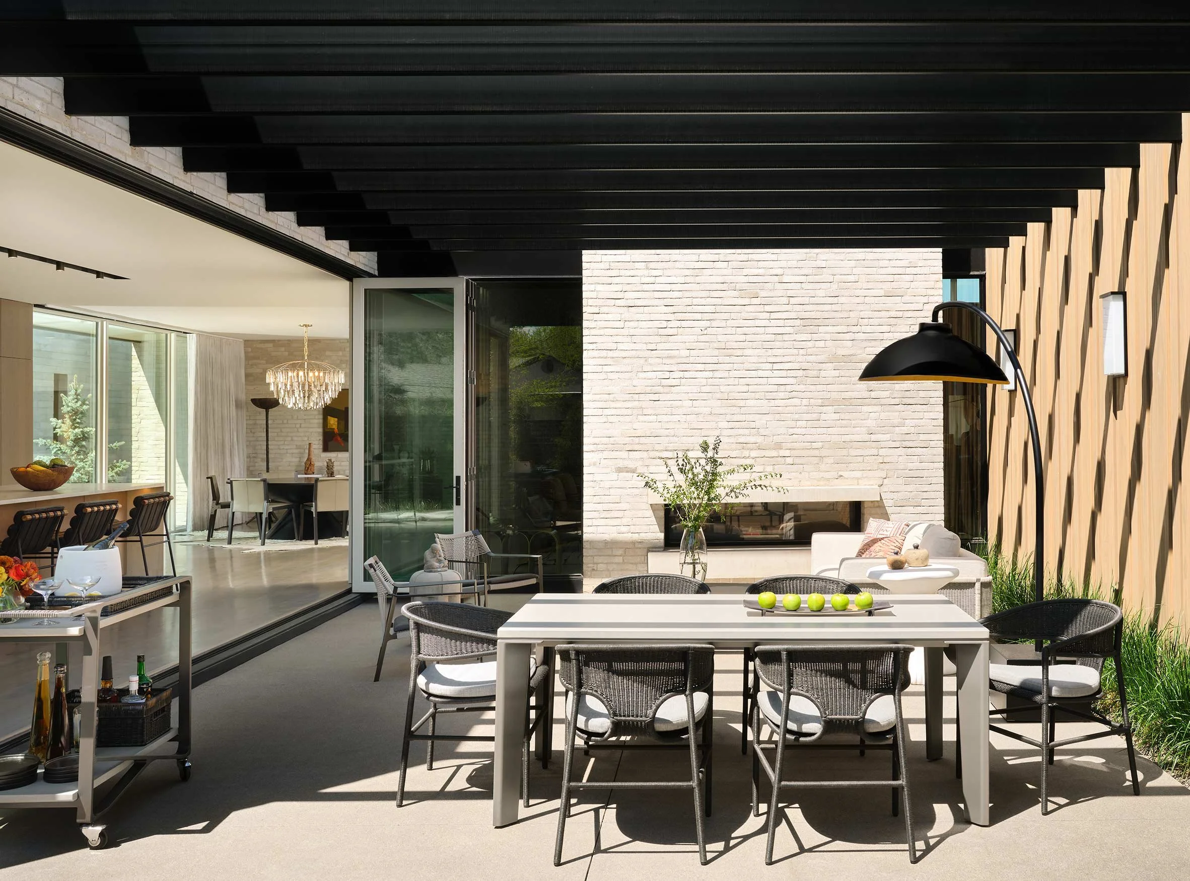 Modern outdoor patio area with dining and seating, large open windows, and a decorative chandelier inside.