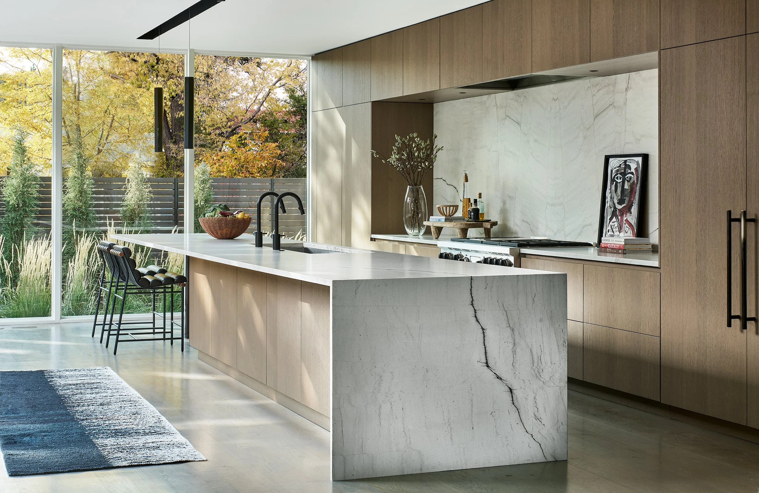 Modern kitchen with large marble island, wooden cabinetry, and large windows showing trees outside with autumn foliage.