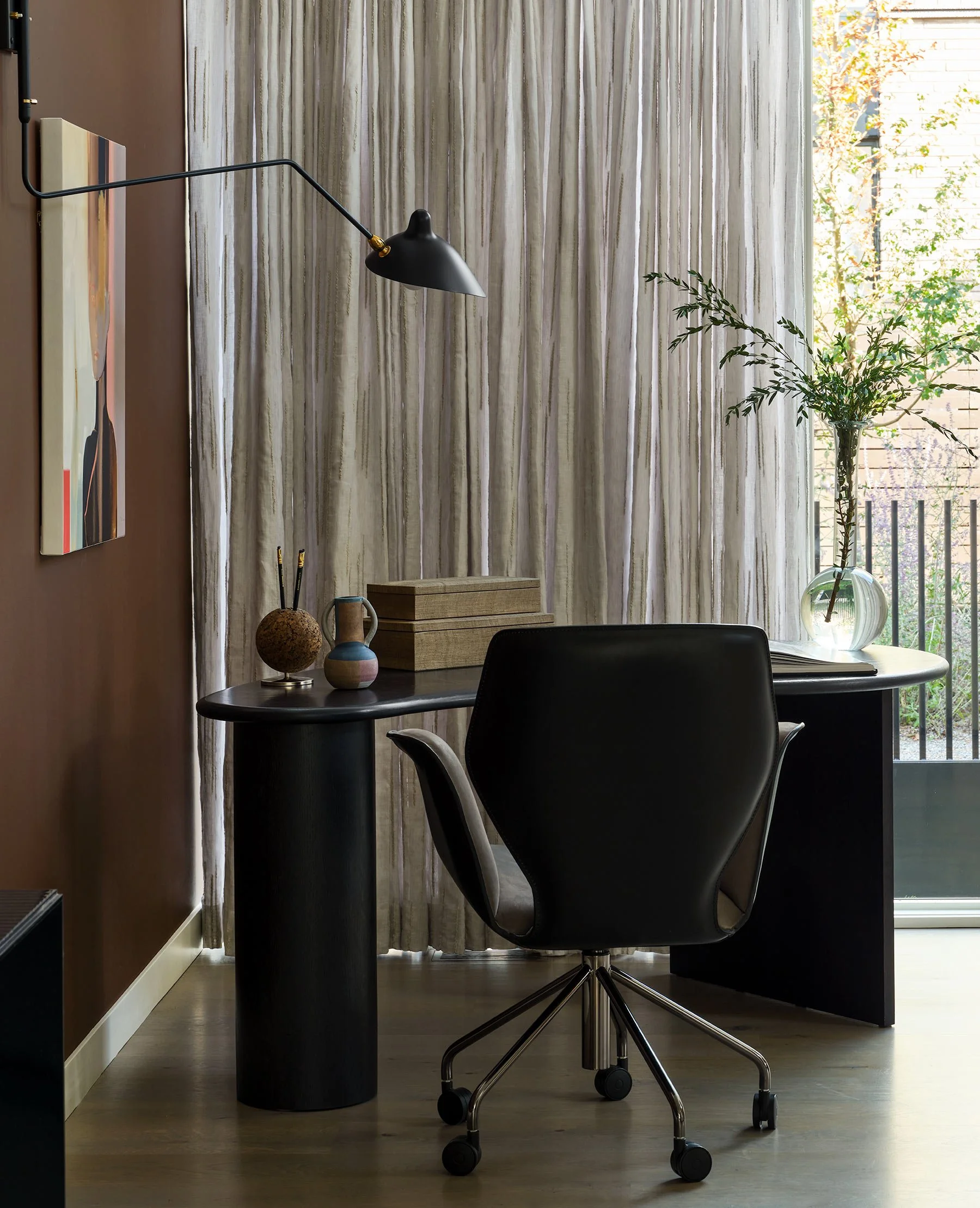 Office space with a black ergonomic office chair, a small round black side table, and a window with curtains. Light coming through the window highlights a vase with green leafy branches, a wooden box, a ceramic mug, and decorative objects on the tabl