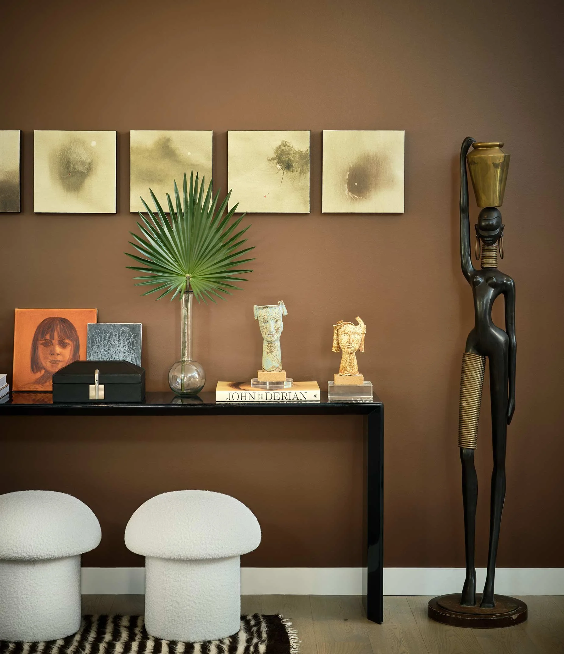 Living room with three white round stools, a black table, a large leafy plant, framed artwork, sculptures, and a tall African-style sculpture against a brown wall.