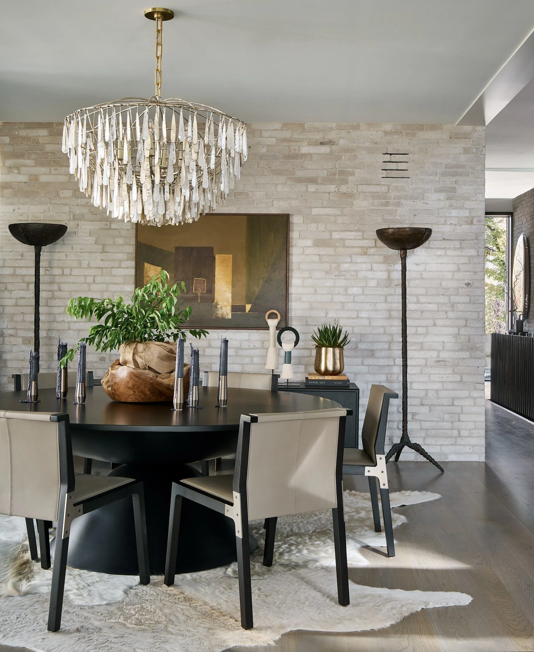 Modern dining room with a dark round dining table surrounded by beige chairs, a large decorative chandelier hanging above, and various decorative objects including candlesticks, plants, and artwork, all set against a light brick wall and natural ligh
