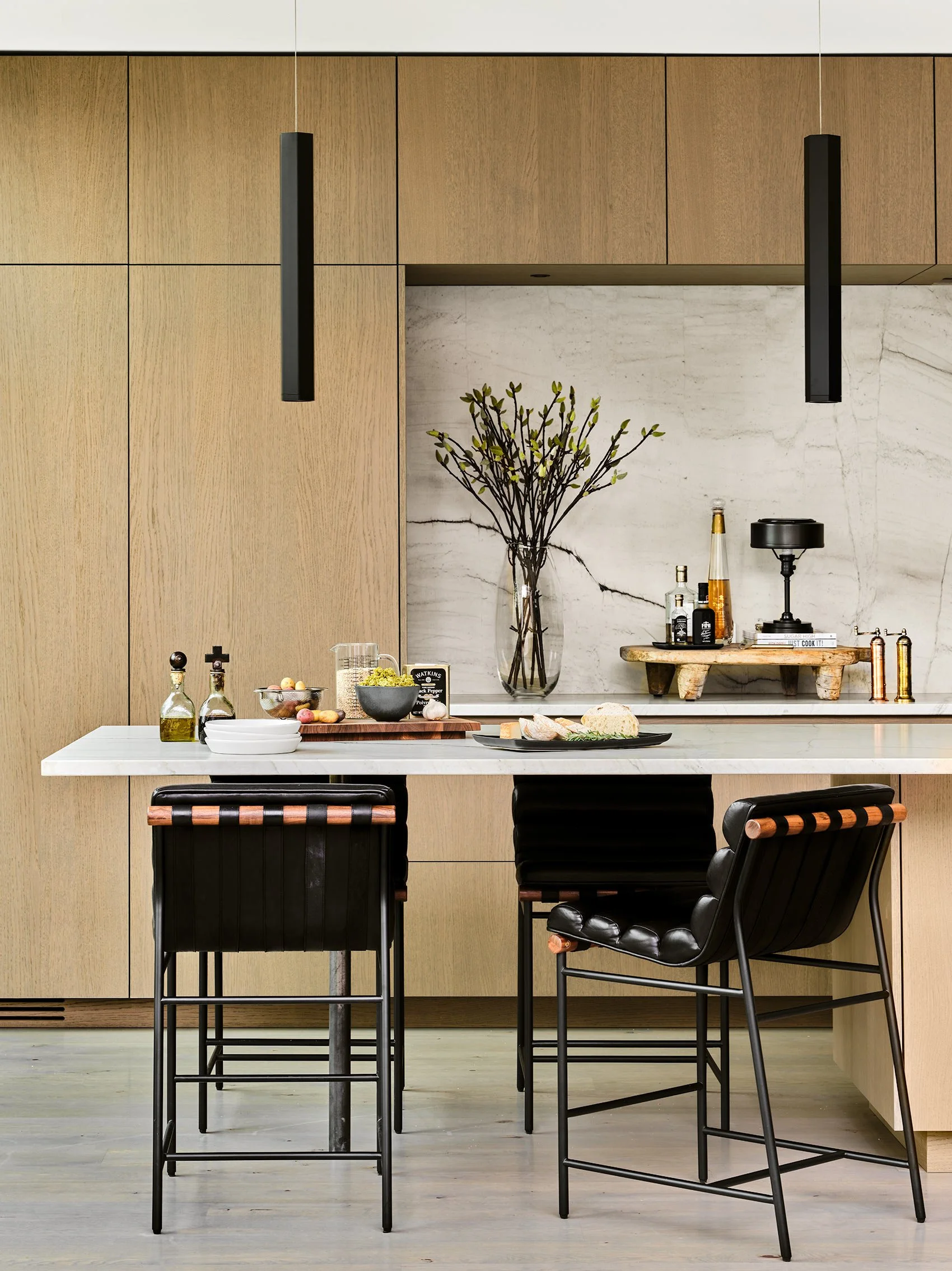 Modern kitchen with wooden cabinets, black hanging pendant lights, and a white marble backsplash. A vase with branches and leaves is placed on the counter, with various bottles and a tray of bread and snacks. Two black chairs are in front of the coun