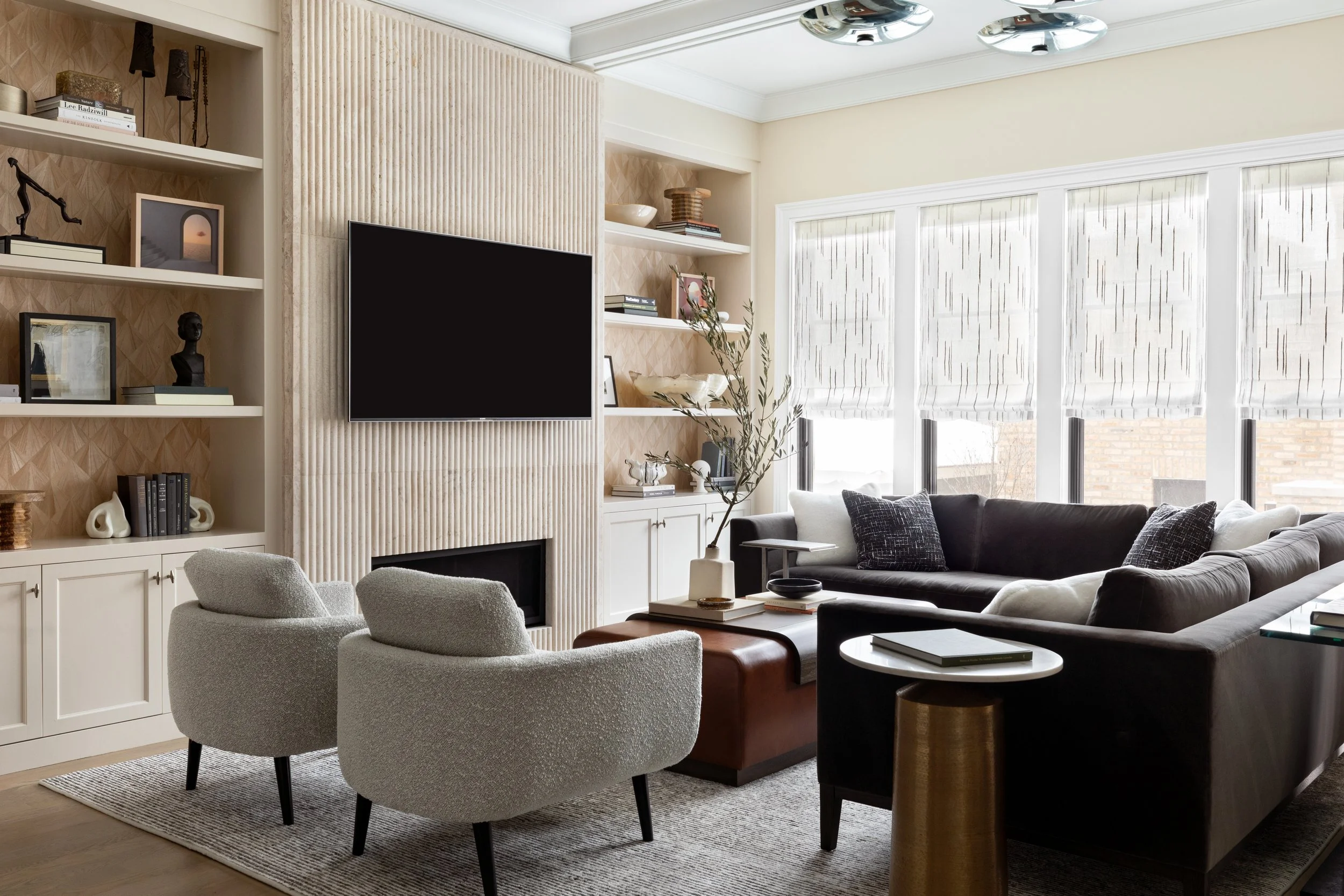 Modern living room with beige walls, a large window with white blinds, a flat-screen TV on the wall, white built-in shelves with decor and books, a gray armchair, a gray sofa with dark pillows, a coffee table, and a beige rug.