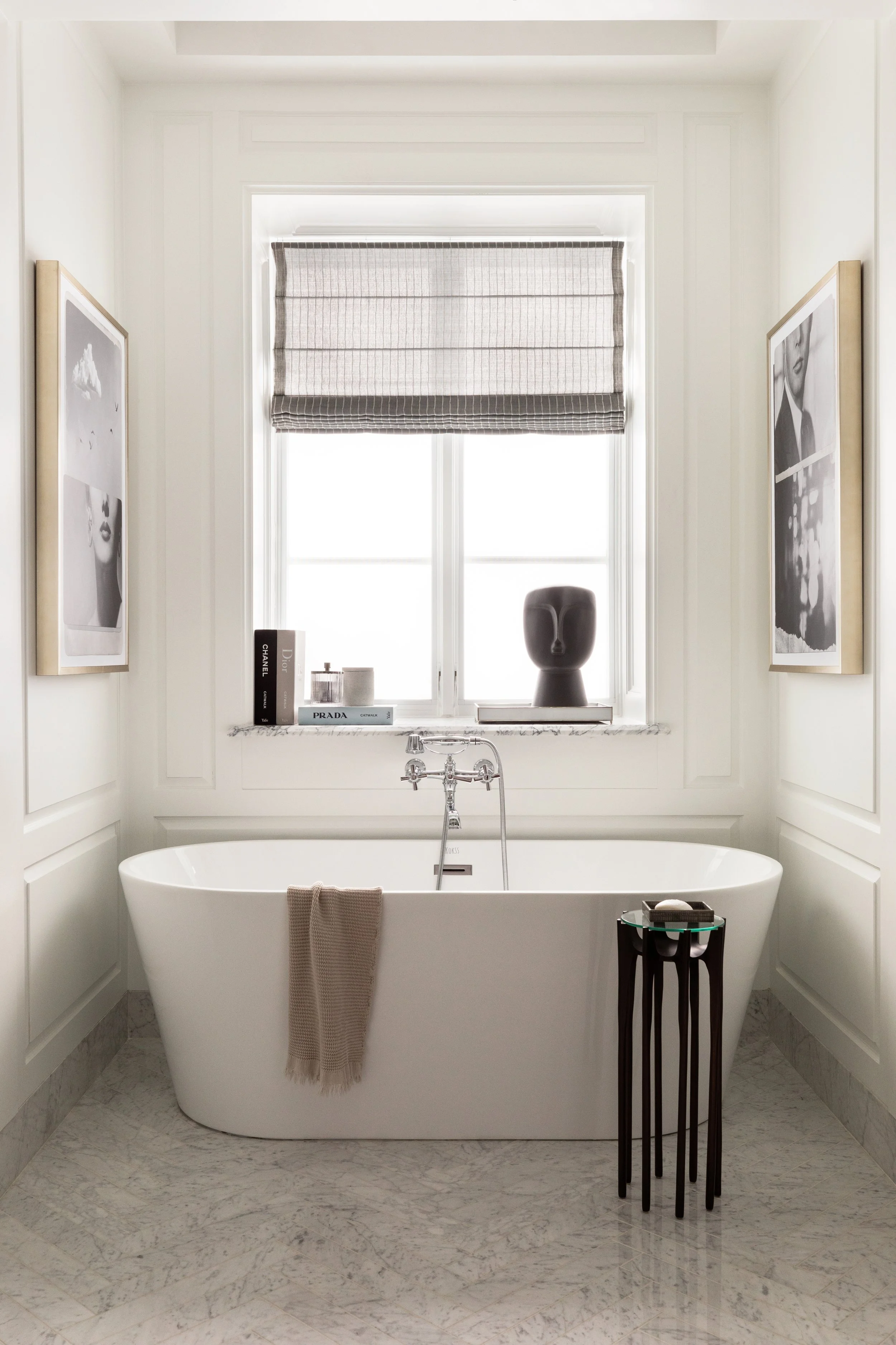 Modern bathroom with a free-standing bathtub, marble floor, and a large window with a Roman shade. Art prints on the walls, a small side table with bath items, and a decorative black vase on the window sill.