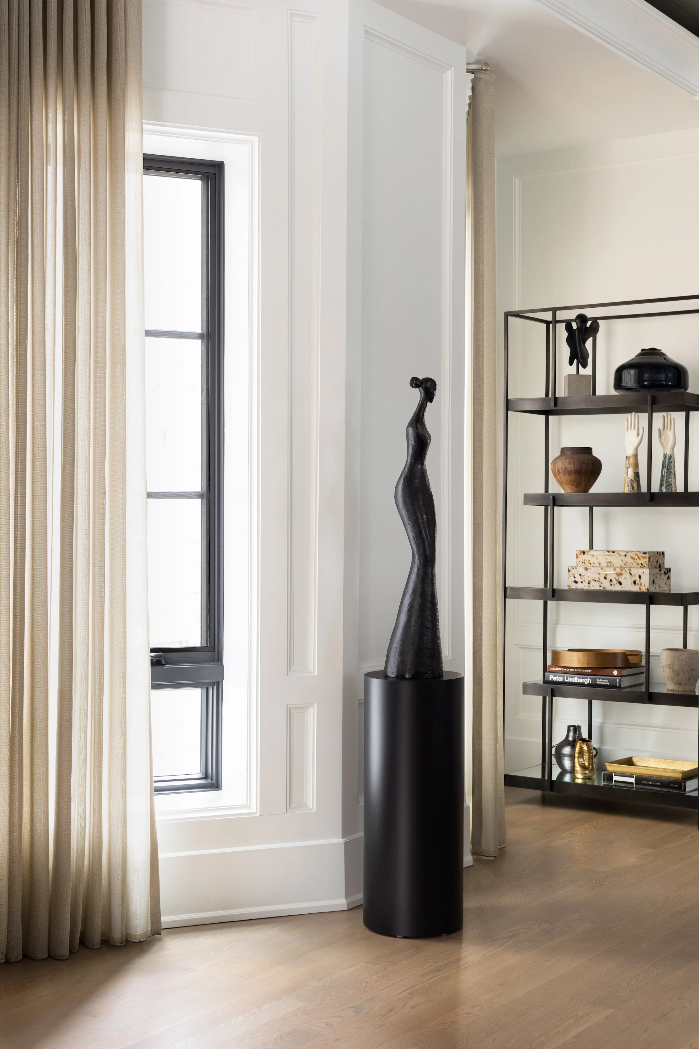 A modern interior space featuring a window with beige curtains, a tall abstract black sculpture on a black pedestal, and a black metal bookshelf with decorative items including vases, books, and art pieces.
