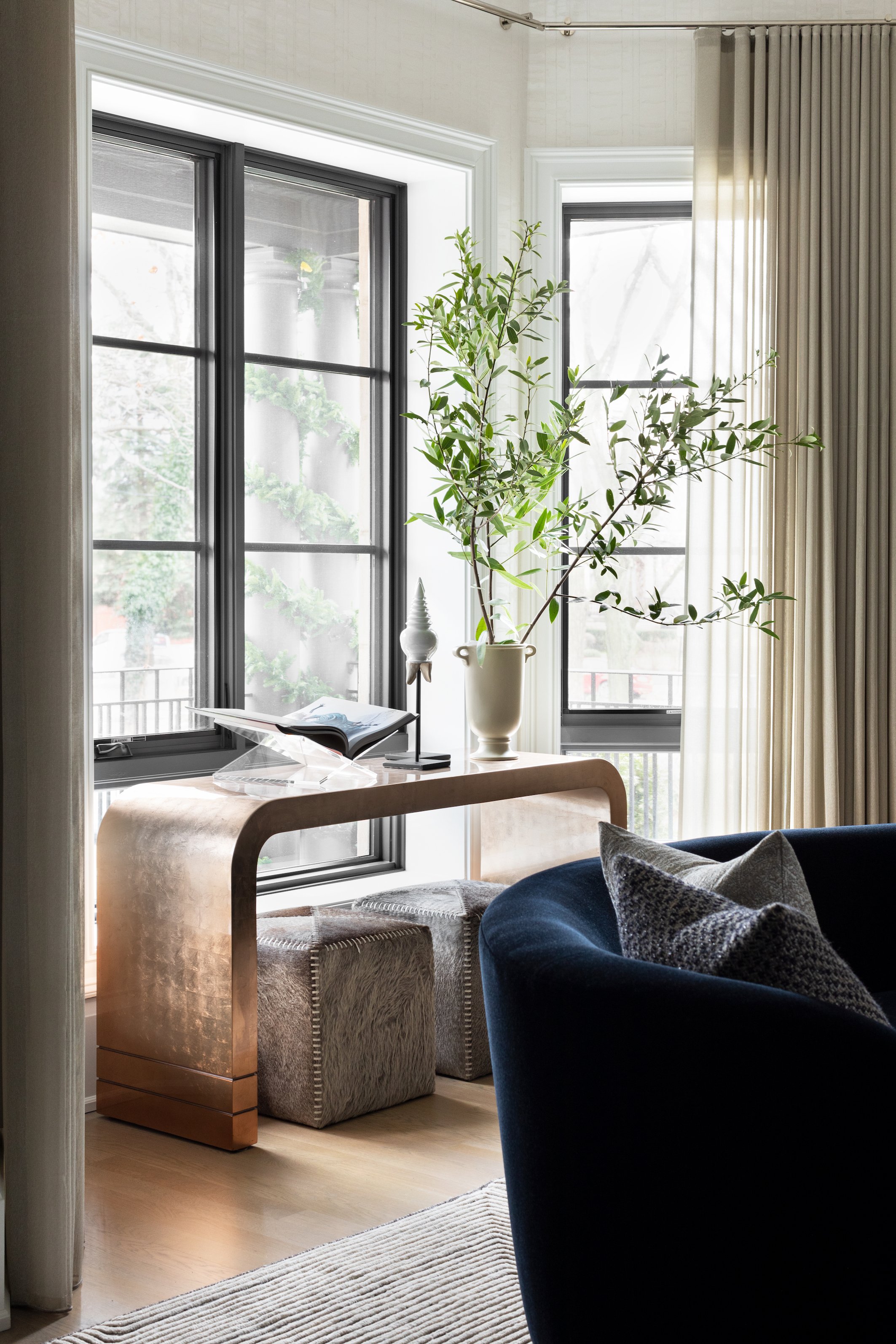 A cozy living room with large windows, a tall potted plant, a velvet blue armchair with pillows, a wooden side table with a book, and beige curtains.